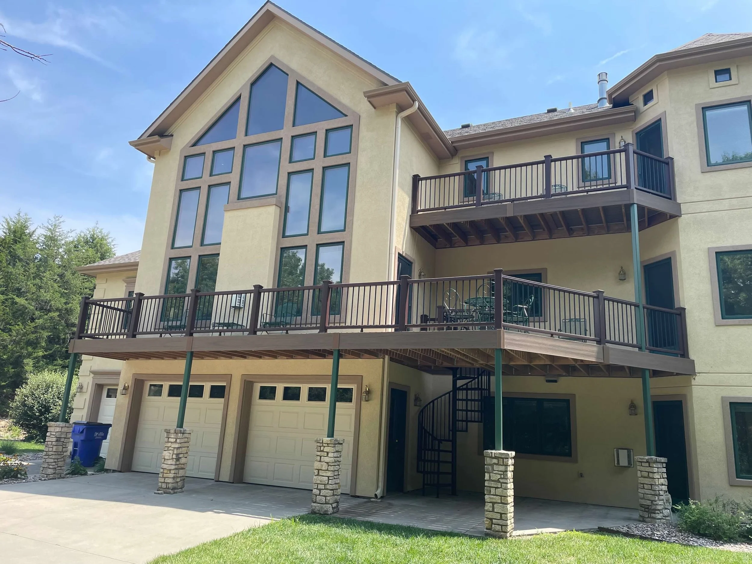 The image shows a multi-story house with beige stucco walls, multiple large windows, and two wooden balconies with black railings. The house has a three-car garage at the ground level, and the roof is gray. There are outdoor furniture and a spiral st
