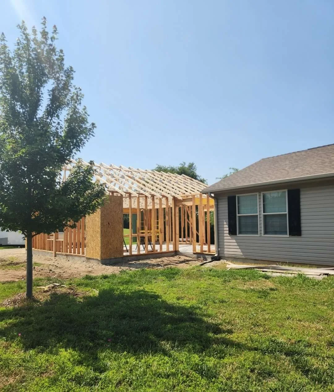 House under construction with a frame and roof trusses, adjacent to an existing house with beige siding and black shutters, in a yard with a tree and green grass.