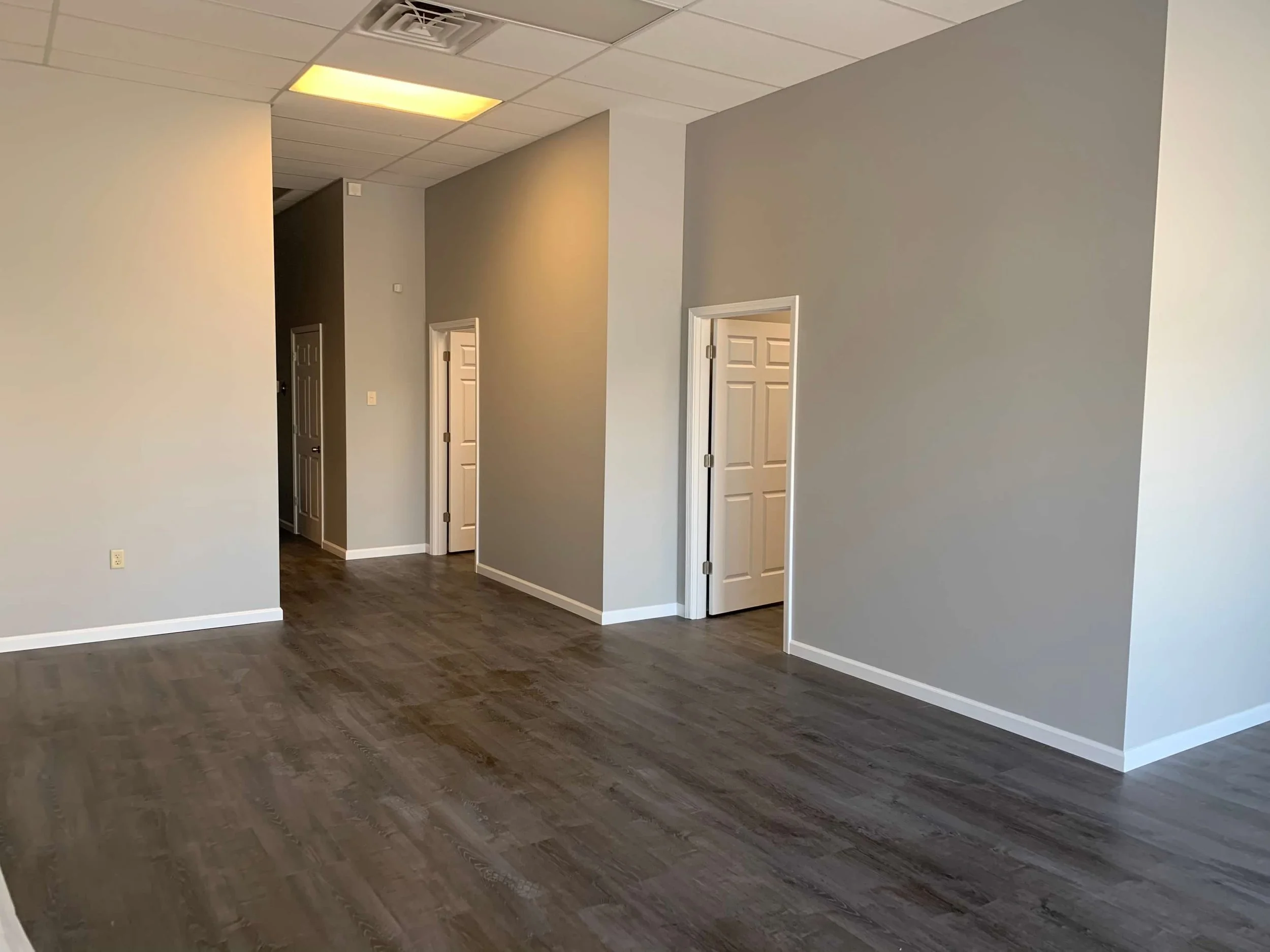 Empty room with gray walls, white baseboards, and dark wood laminate flooring. There are three white doors, one partially open, and a ceiling with recessed lighting and a fluorescent light panel.