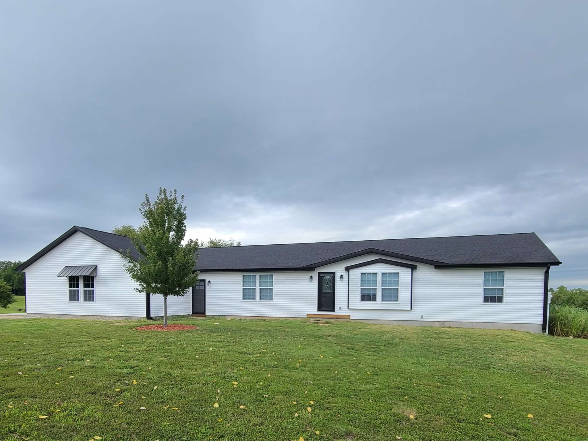 Single-story white house with black roof and shutters, front door with small stairs, surrounded by green lawn and a tree, overcast sky.