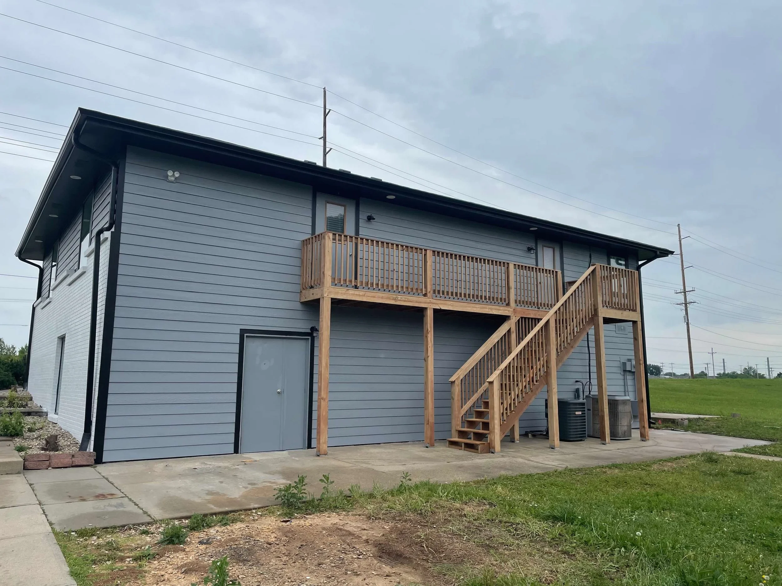 A two-story gray house with black trim, featuring a wooden staircase and balcony on the second floor. The house has a concrete patio and some greenery in the yard, with utility meters and HVAC units outside.