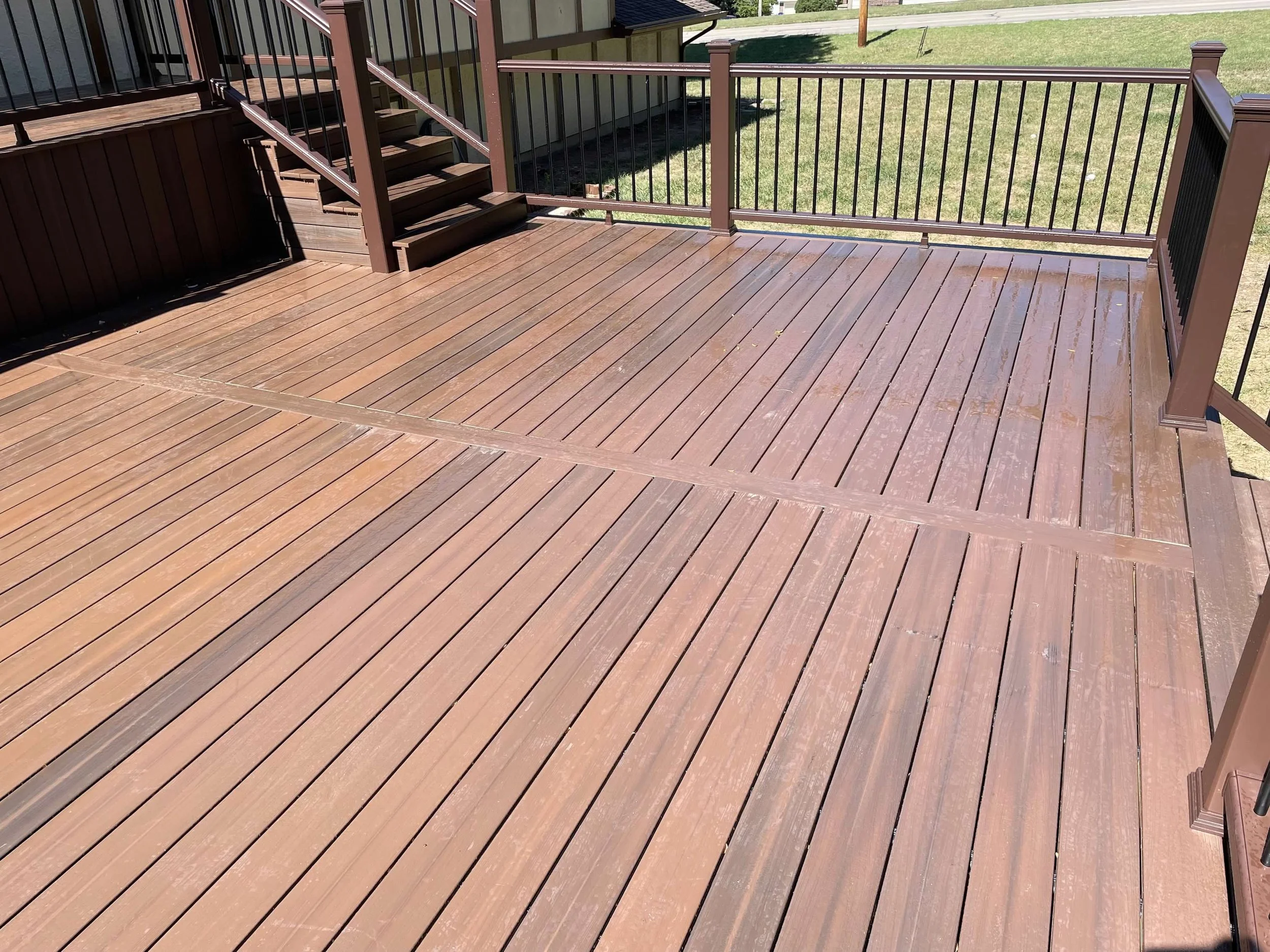 Wooden deck with a brown railing and stairs, showing sunlight and shadow patterns on the surface, with grass visible in the background.