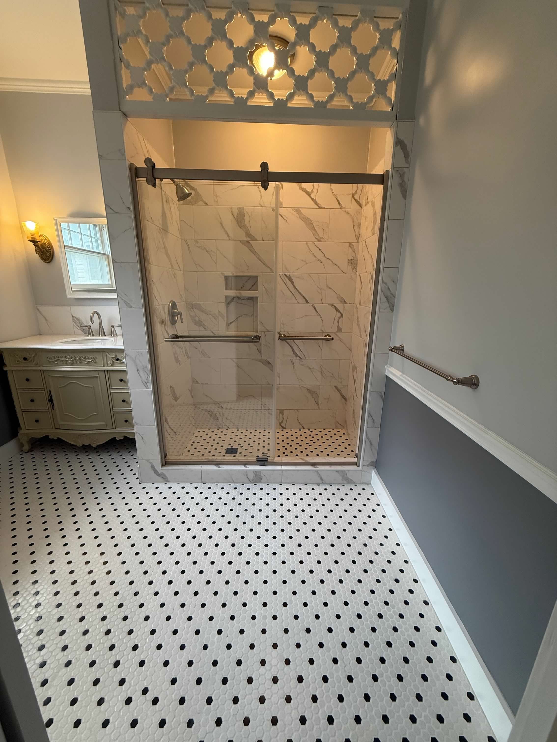 Bathroom with black and white hexagonal tile floor, shower with marble tiles and glass door, vintage vanity with sink, wall-mounted light fixtures, and a window.