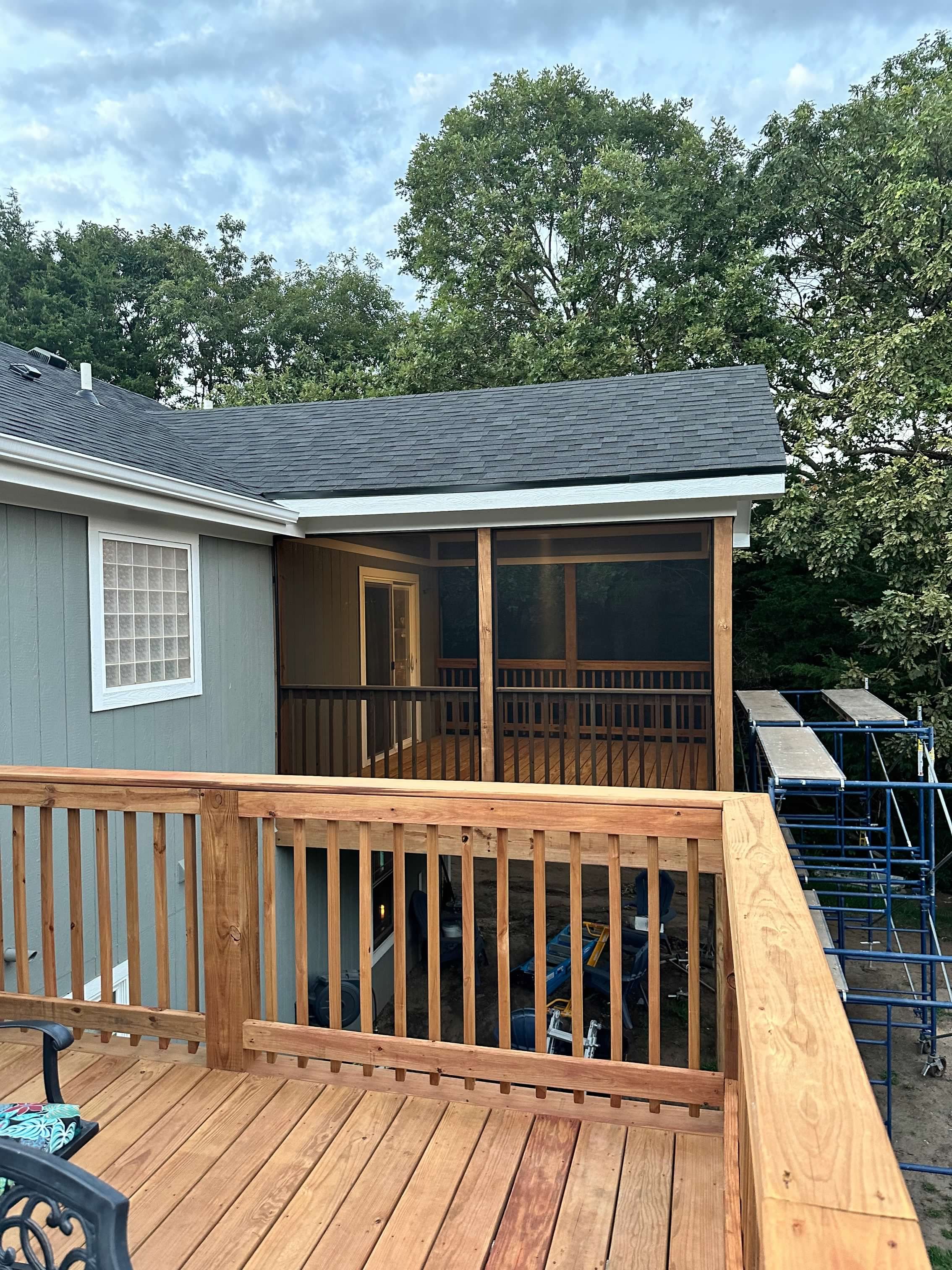 Newly constructed wooden deck and screened porch attached to a house, with some construction equipment visible underneath, surrounded by trees under a partly cloudy sky.
