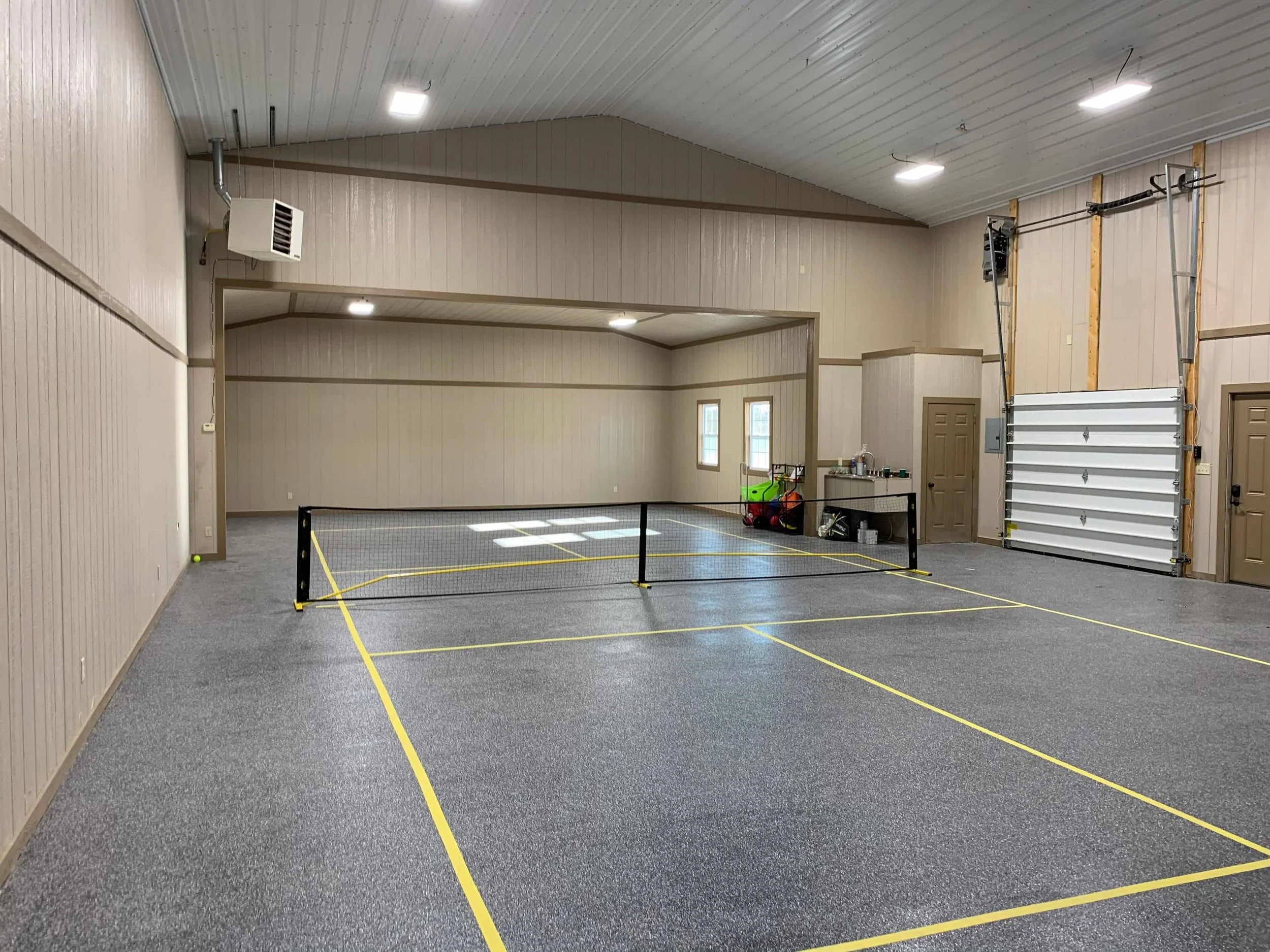 Indoor space with a small tennis court marked with yellow lines, a net in the center, and a garage door on the right side. The room has wood-paneled walls, high ceilings, and ceiling lights.