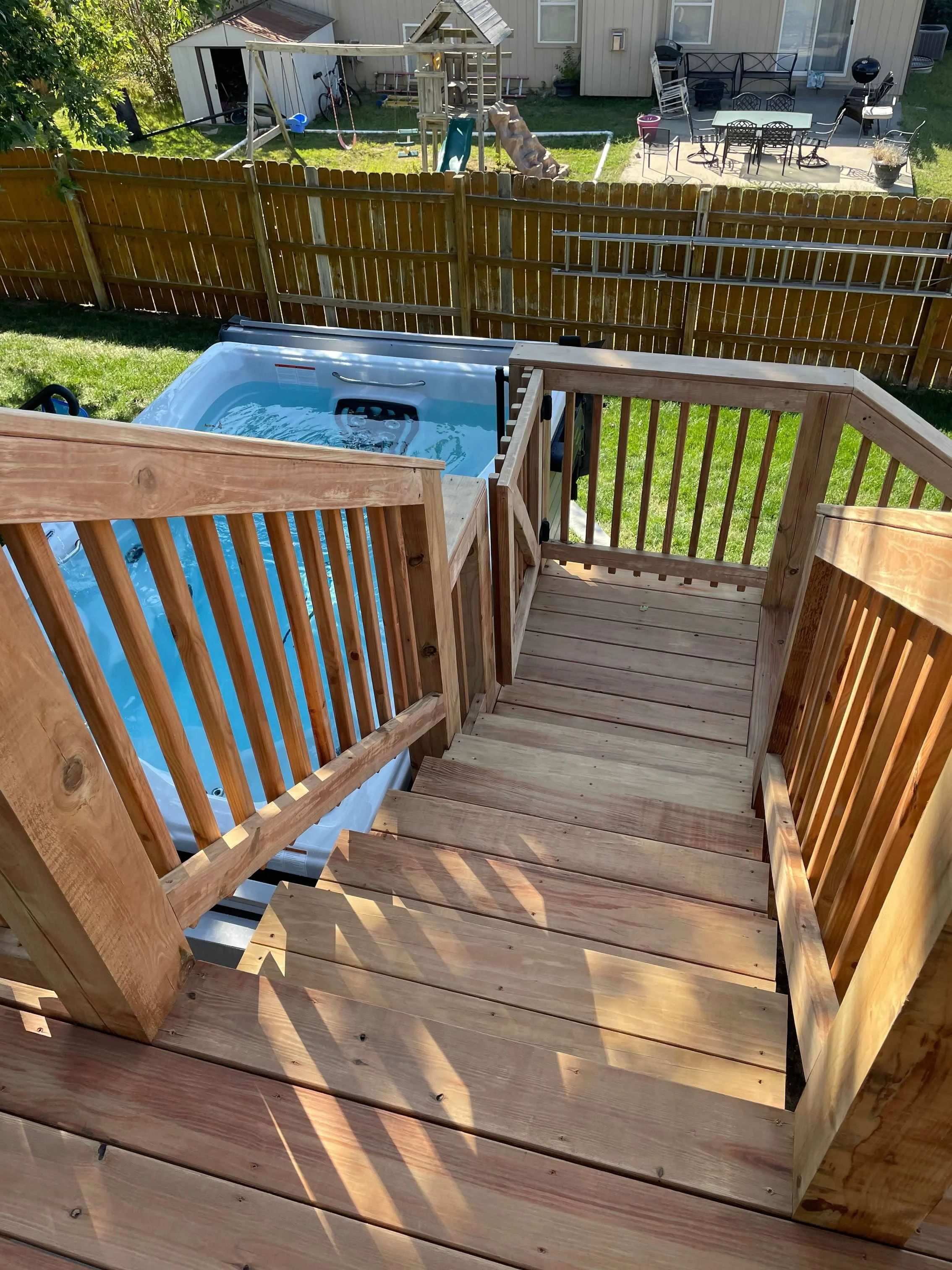 Wooden deck stairs leading down to a backyard with a hot tub, fenced yard, and backyard patio with outdoor furniture.