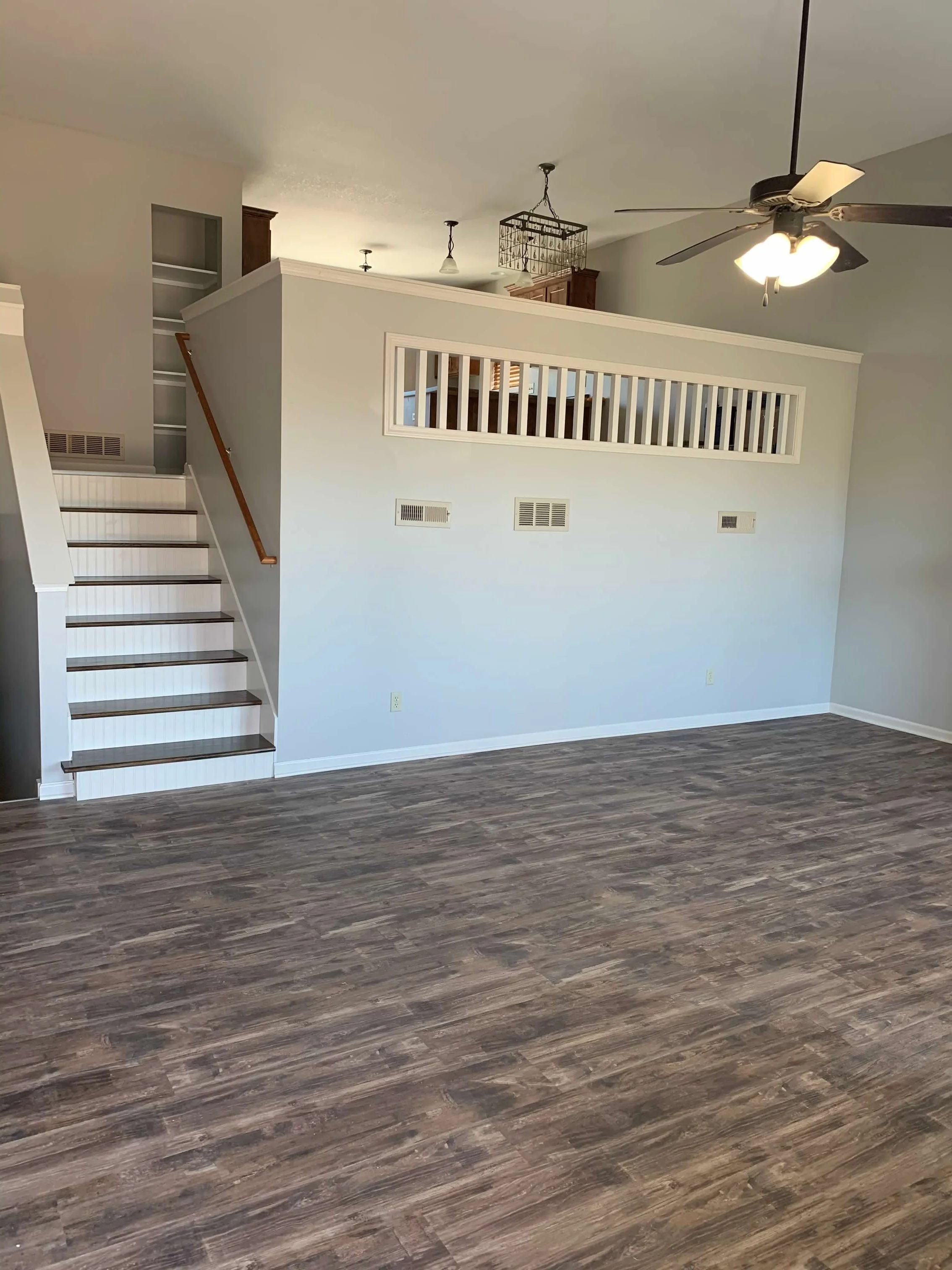 Empty living room with wood flooring, white walls, a ceiling fan, and a staircase leading to a second floor with an open railing.