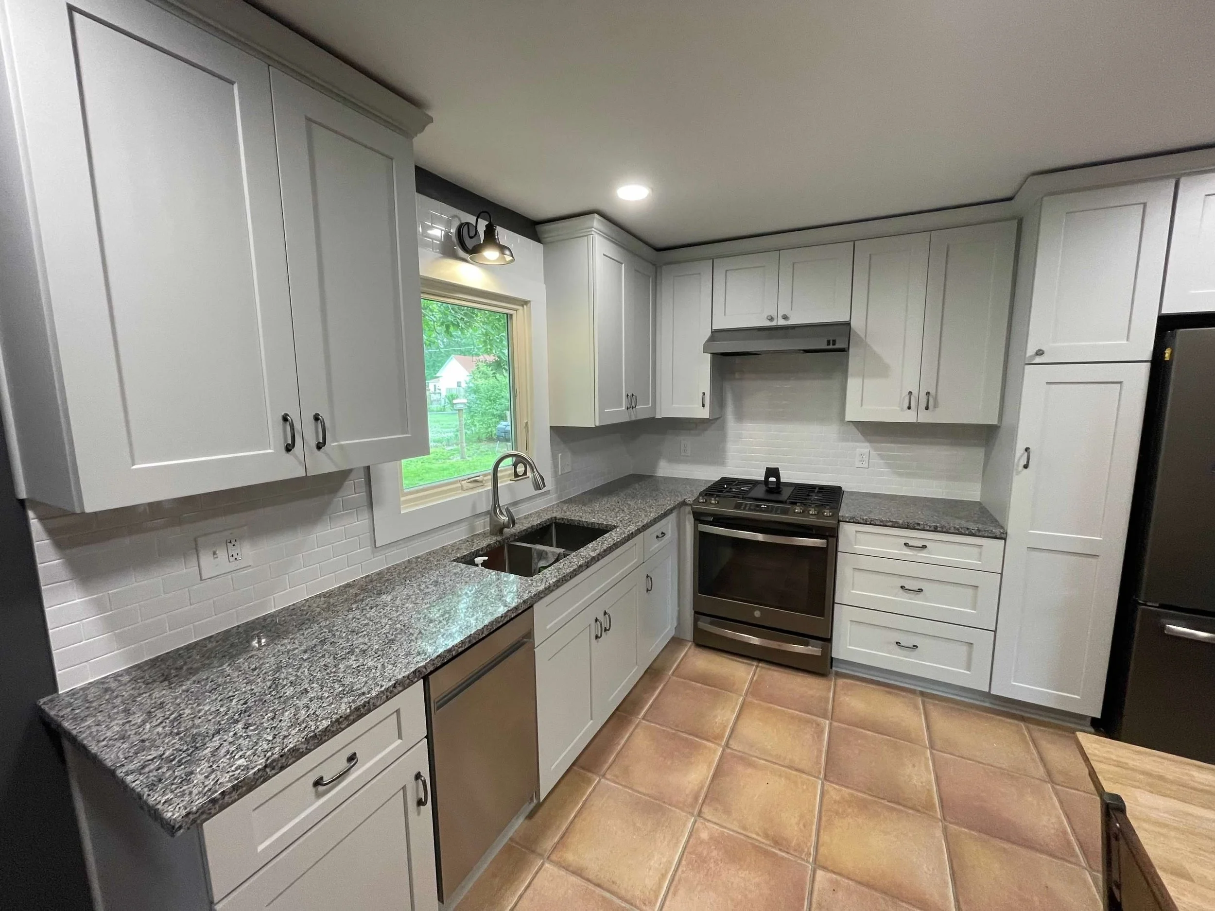 A kitchen with white cabinets, gray granite countertops, a stainless steel stove and dishwasher, a window above the sink, and terracotta tile flooring.