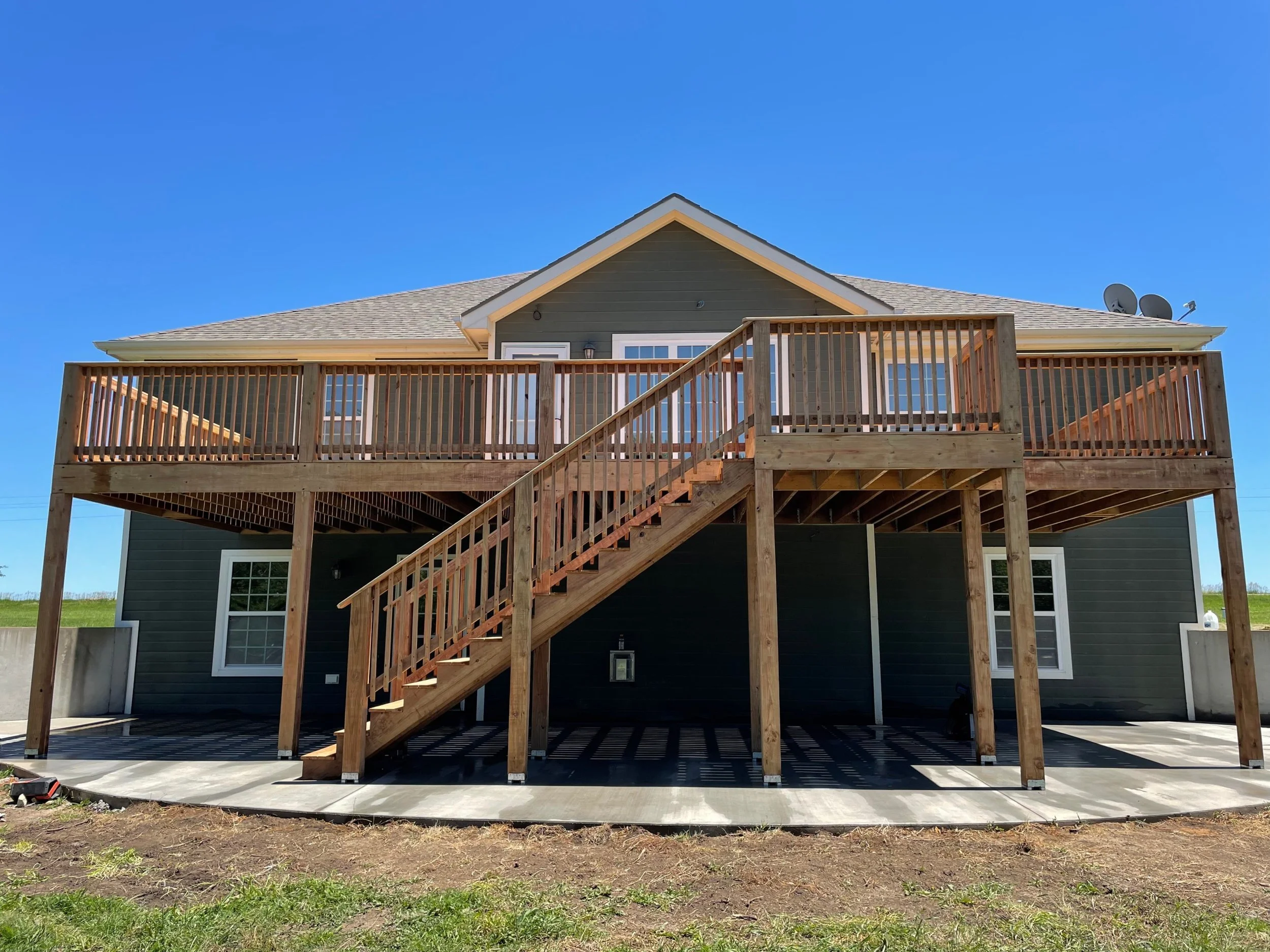 Rear view of a two-story house with a newly built wooden deck and stairs, with satellite dishes on the roof, under a clear blue sky.