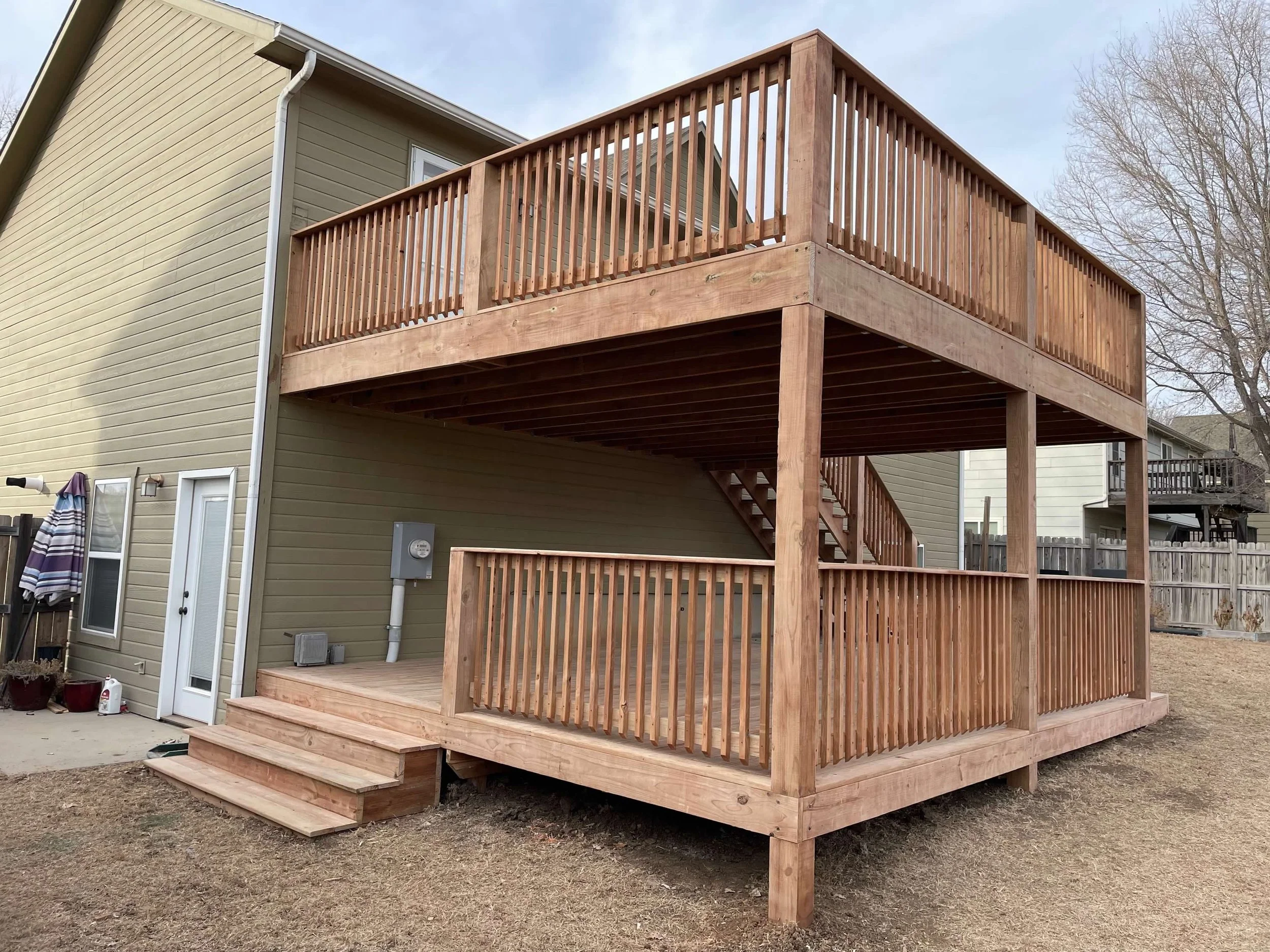 A newly built two-story wooden deck with stairs in the backyard of a house, with a beige siding exterior and a fenced yard.