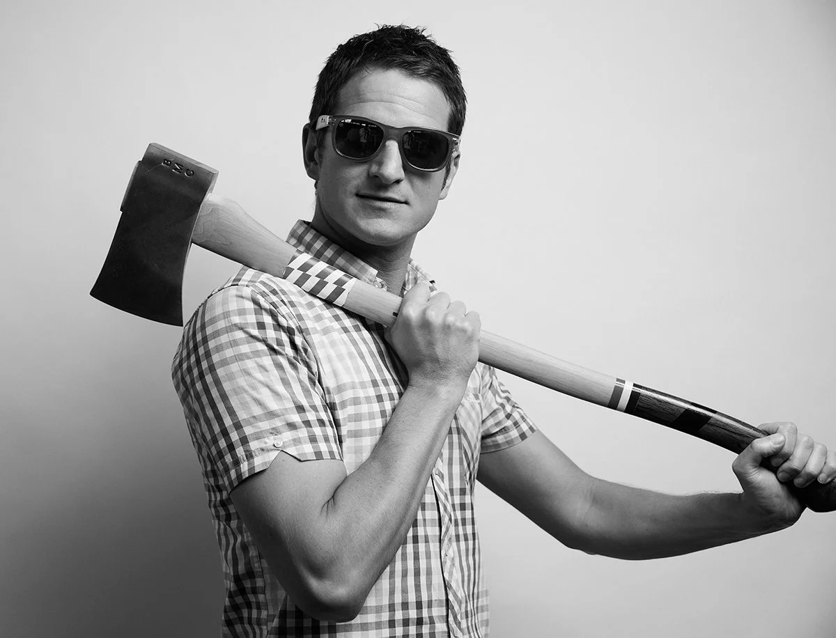 Black and white photo of a man wearing sunglasses and a checkered shirt, holding an axe over his shoulder with one hand against a plain background.