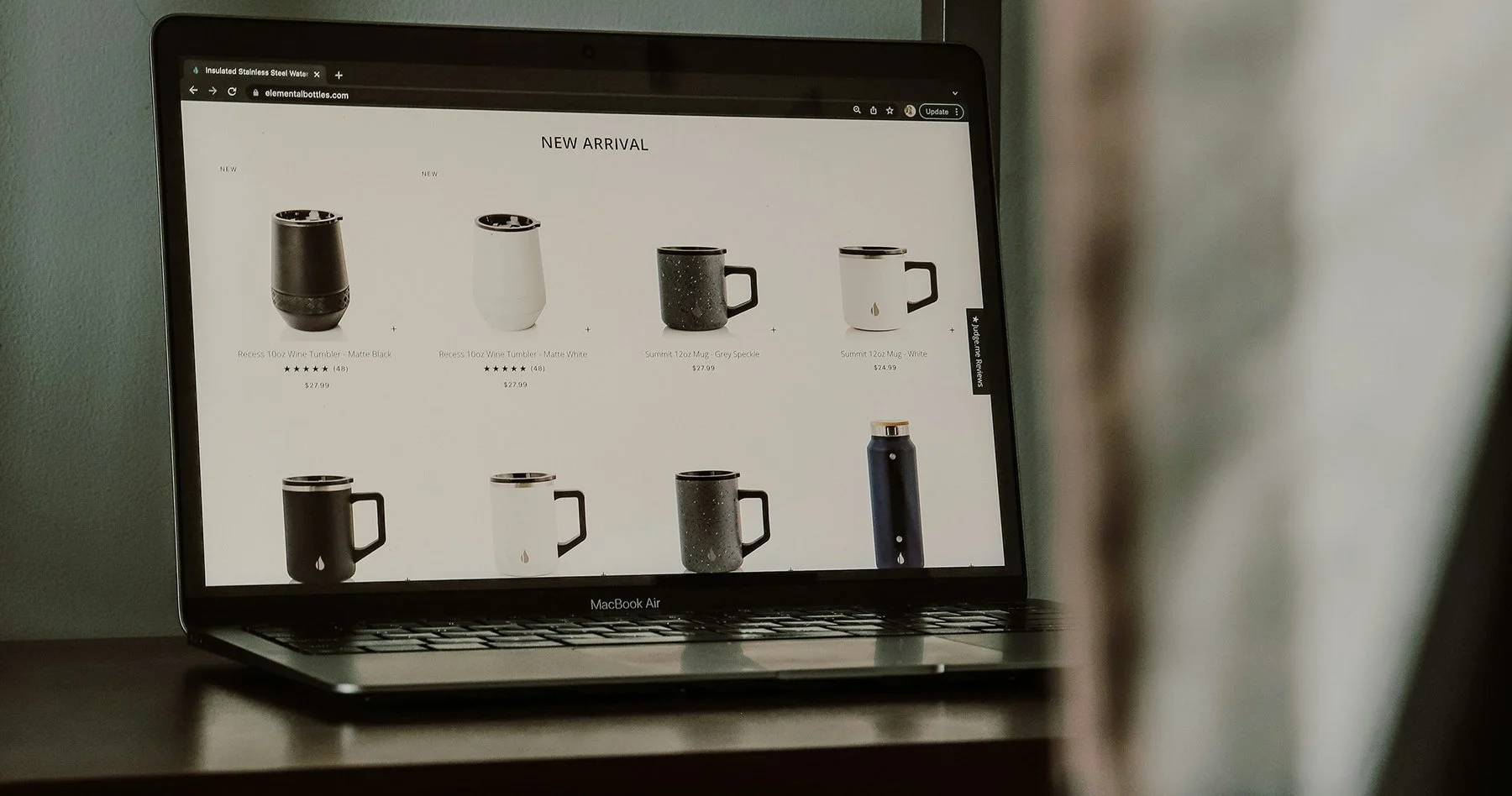 Laptop screen displaying an online shop page featuring ceramic mugs and tumblers labeled as new arrivals, with prices and star ratings.