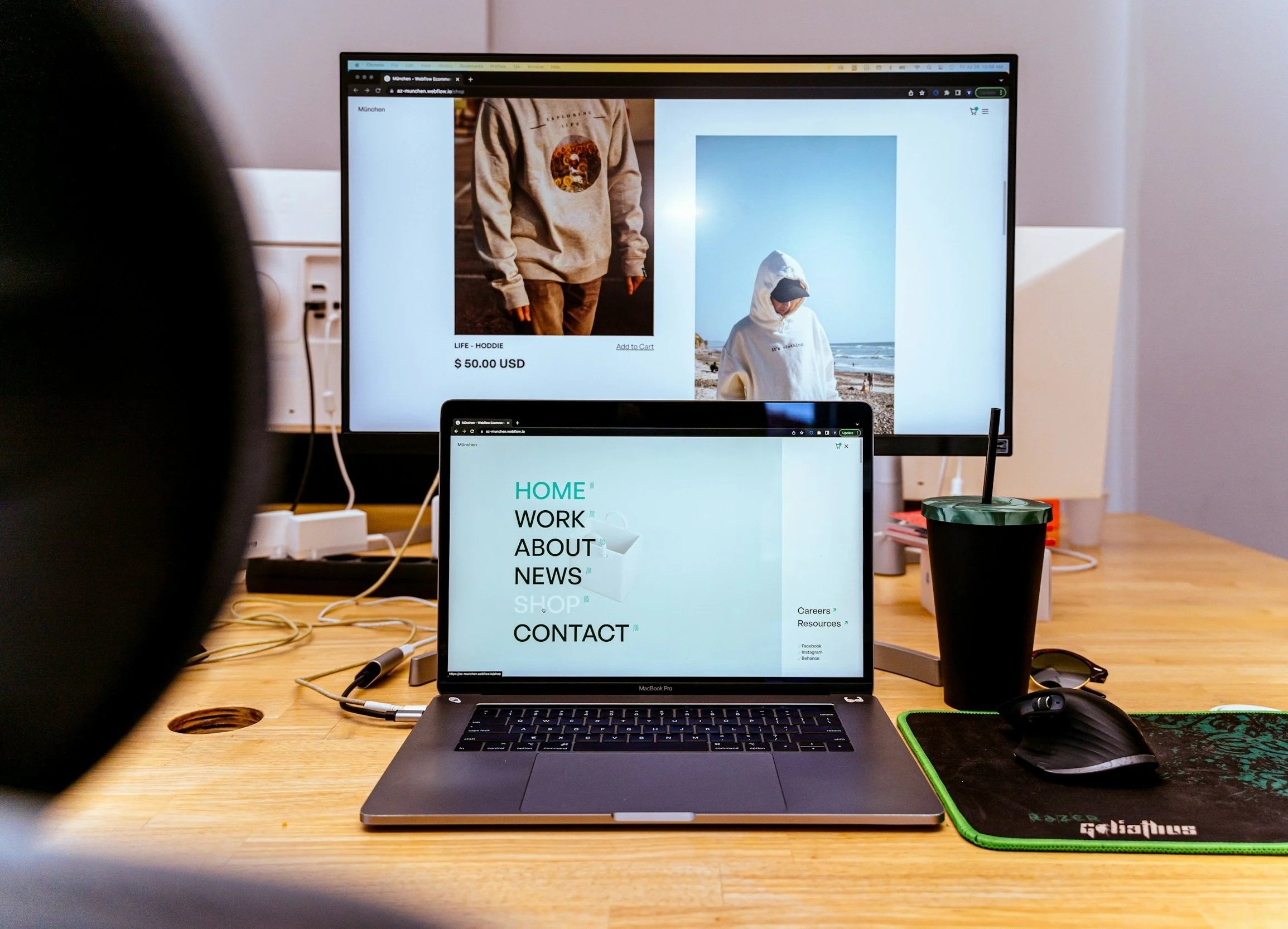 Desk with a MacBook Pro laptop, a black and green mouse pad, a pen, a container with a drink, and a large monitor displaying a website with images of hoodies and a menu with sections for Home, Work, About, News, Shop, and Contact.