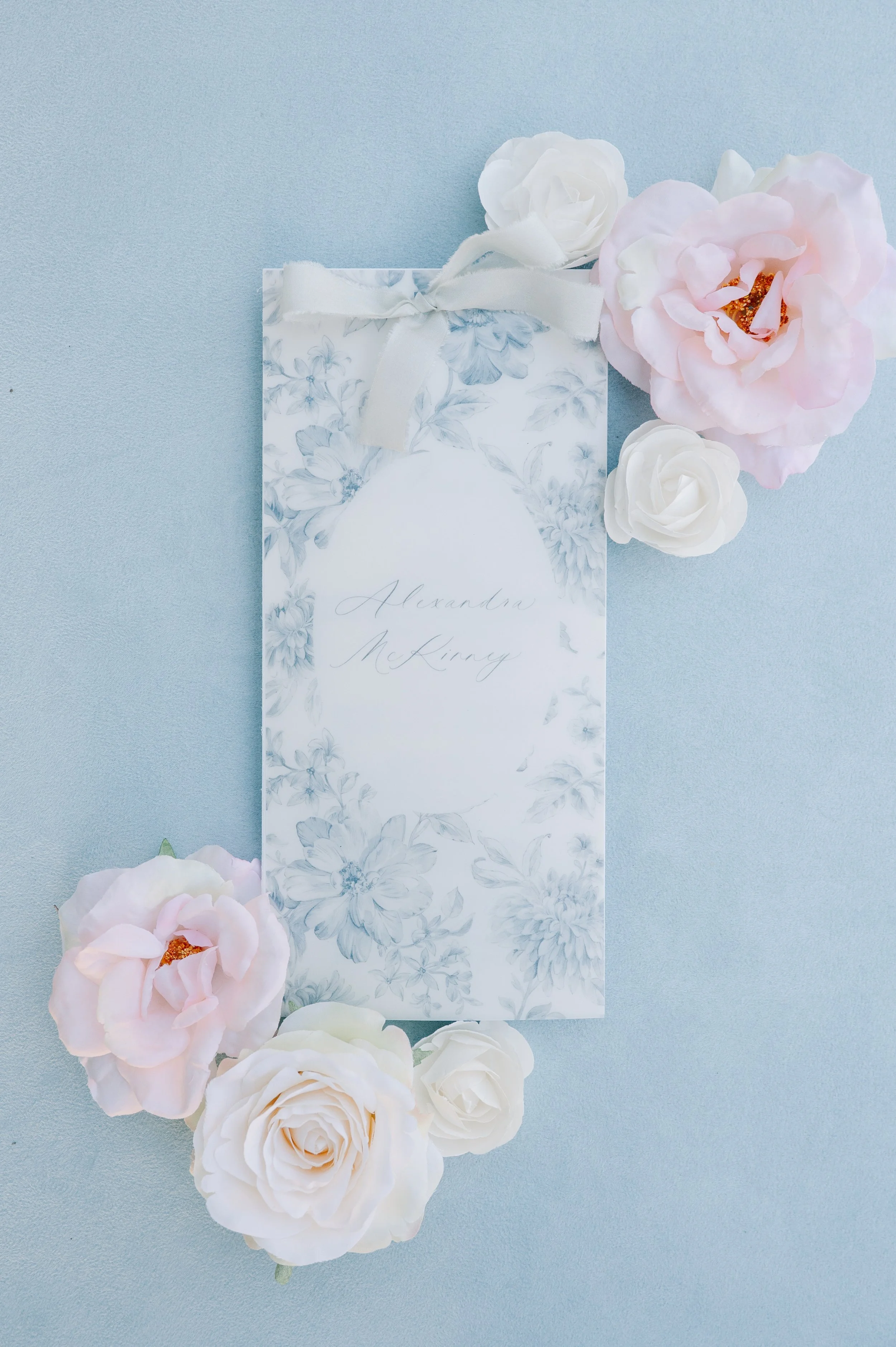 Invitation card with floral design and cursive writing, surrounded by pink and white roses on a pale blue background.