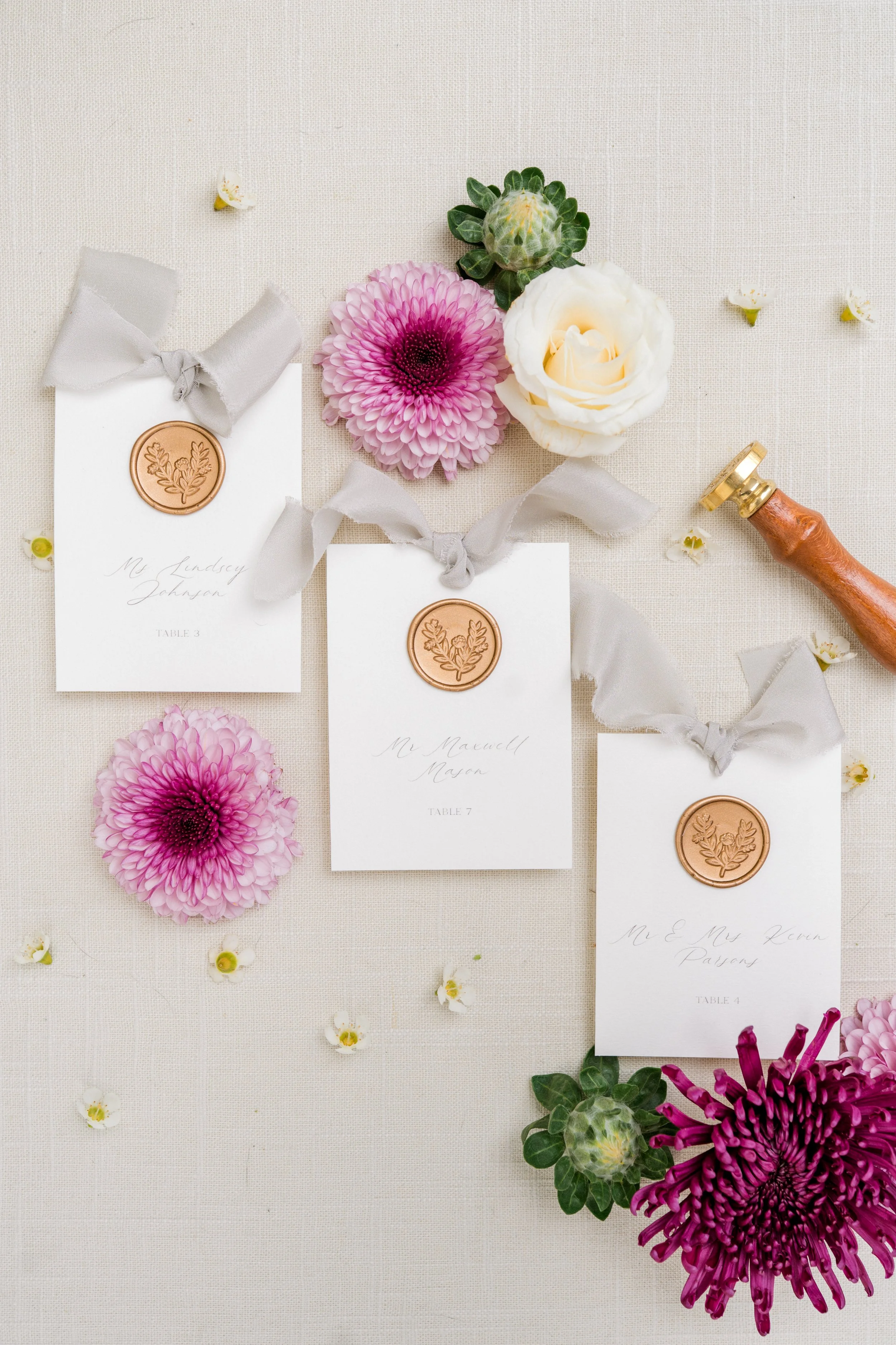 Wedding place cards with gray ribbons and copper wax seals, pink and white flowers, scattered small white flowers, and a wax stamp on a beige fabric background.