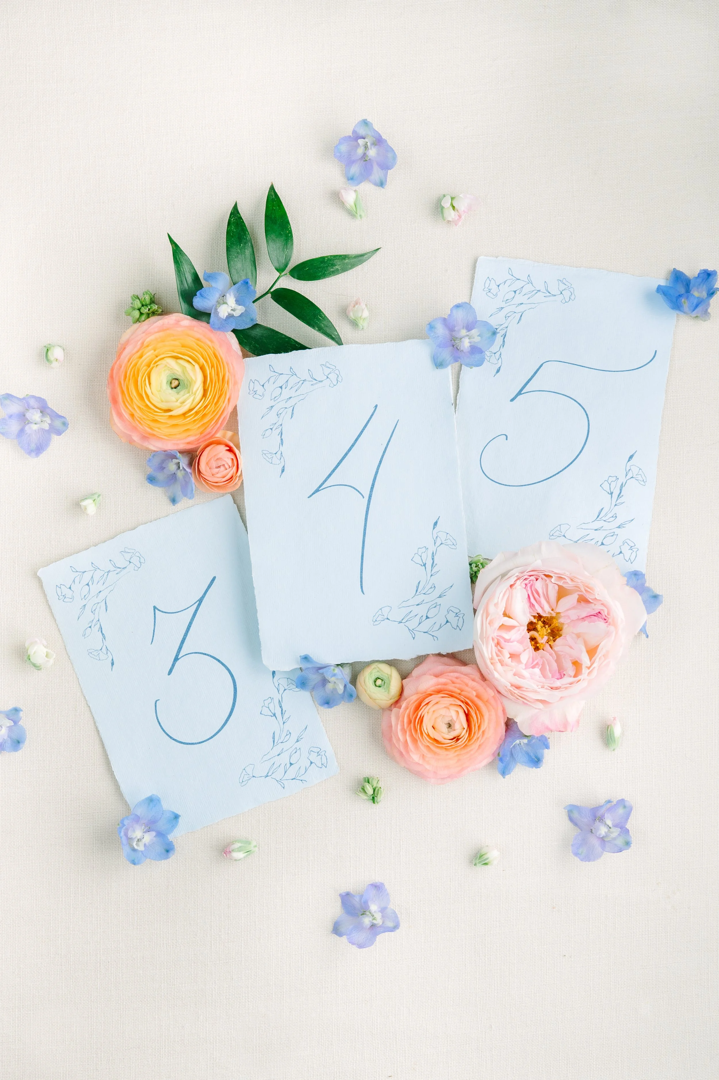 Three pieces of blue paper with handwritten numbers 3, 4, and 5, surrounded by pink and peach roses, blue flowers, green leaves, and scattered small flower petals on a light-colored background.
