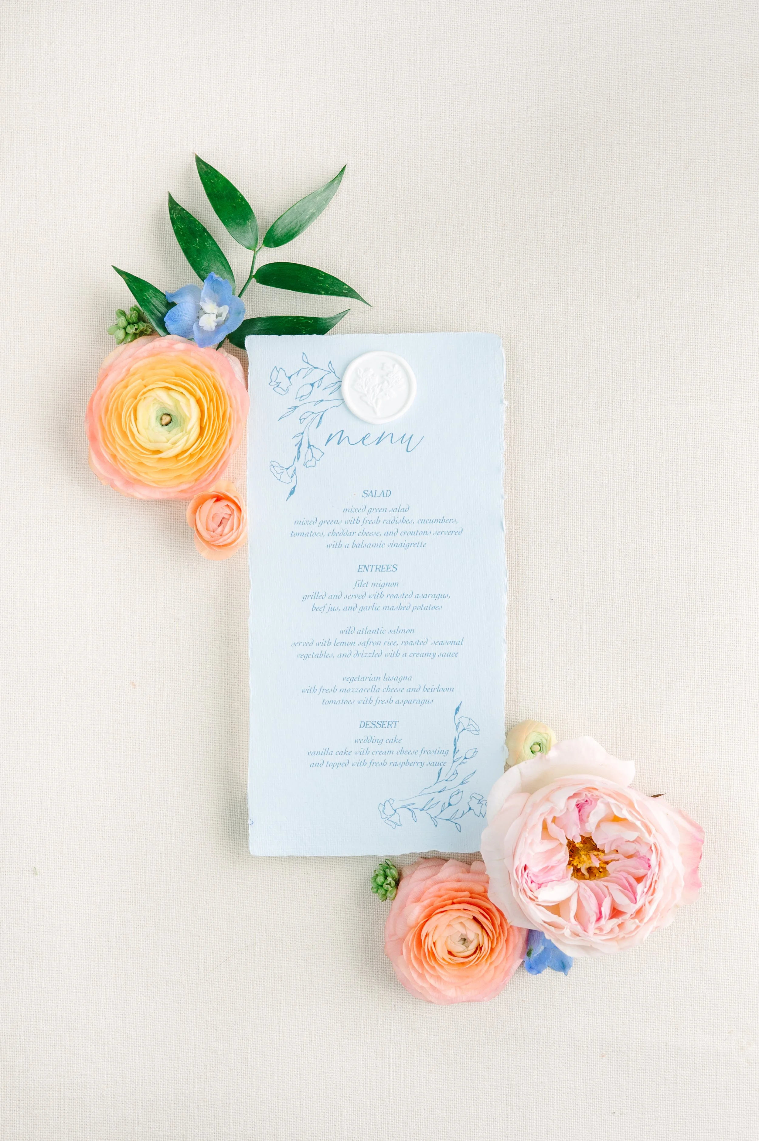 Wedding menu on light blue paper with floral illustrations, surrounded by pink, peach, and cream roses, green leaves, and small blue flowers on a light fabric background.