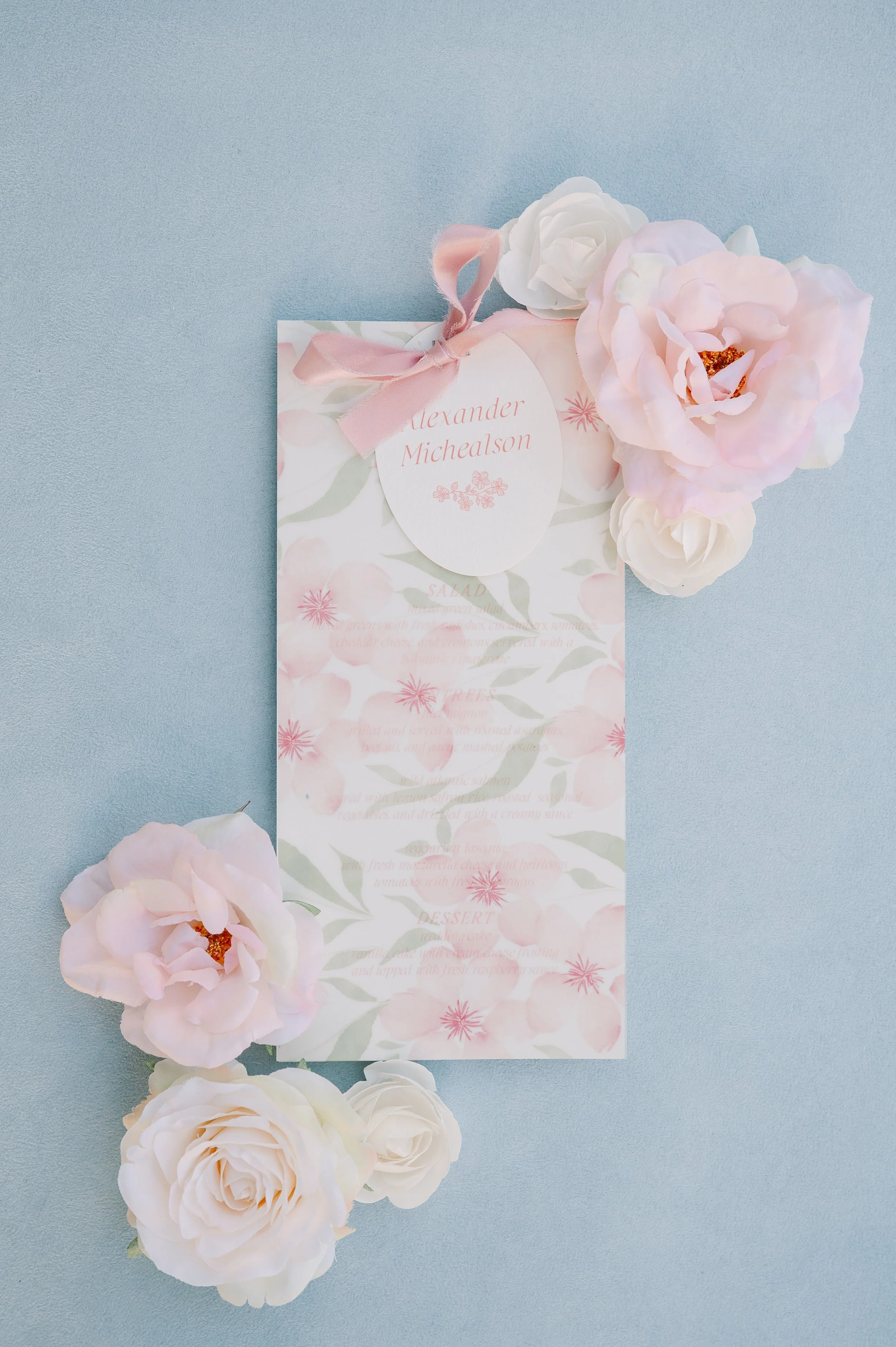 A pale pink and white flower arrangement around a floral-themed menu card with a pink ribbon and a round tag that reads 'Alexander Michealson'.