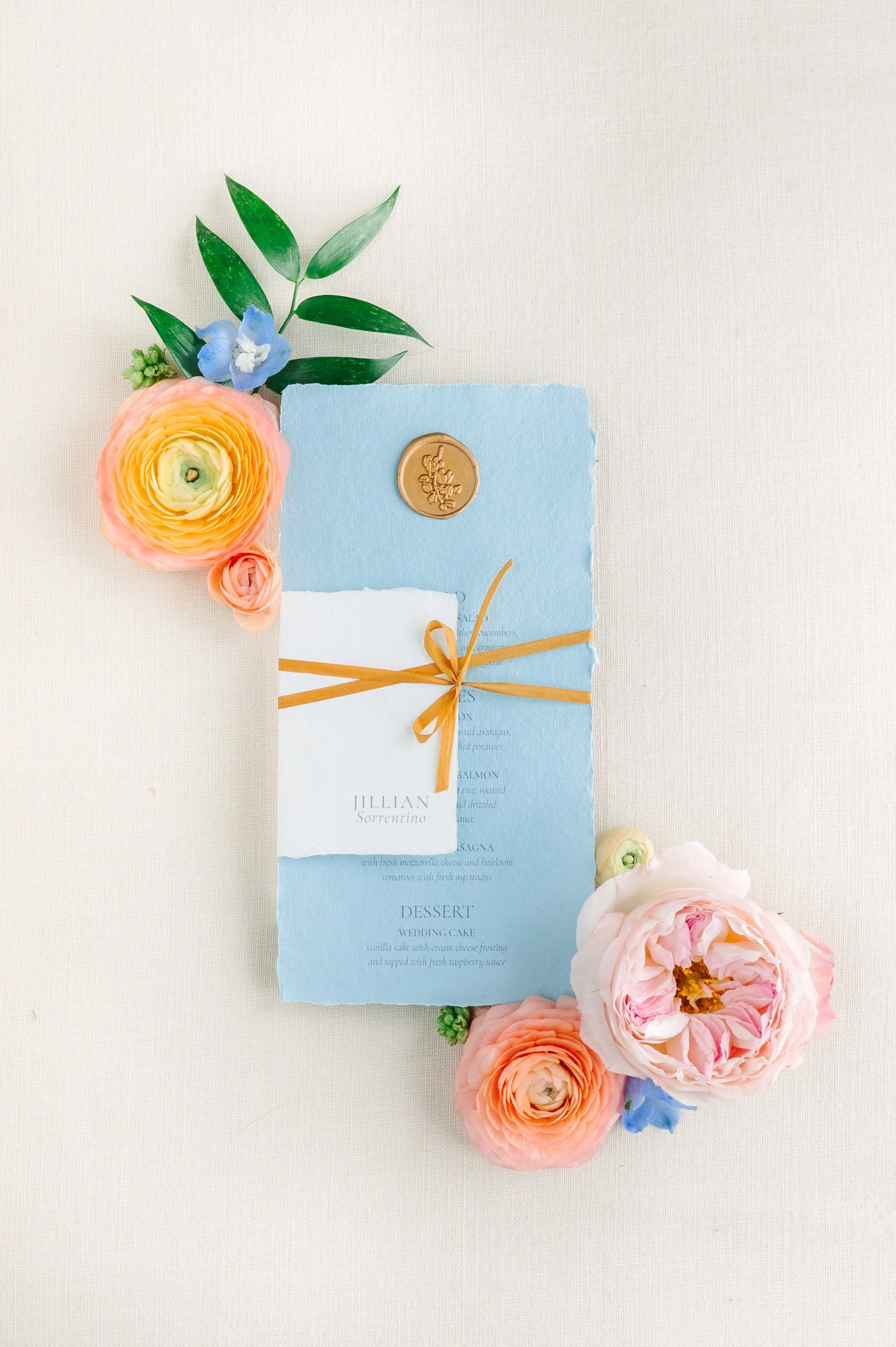 A light blue wedding menu card decorated with a gold ribbon and sealing wax, surrounded by pink, peach, and blue flowers on a neutral background.