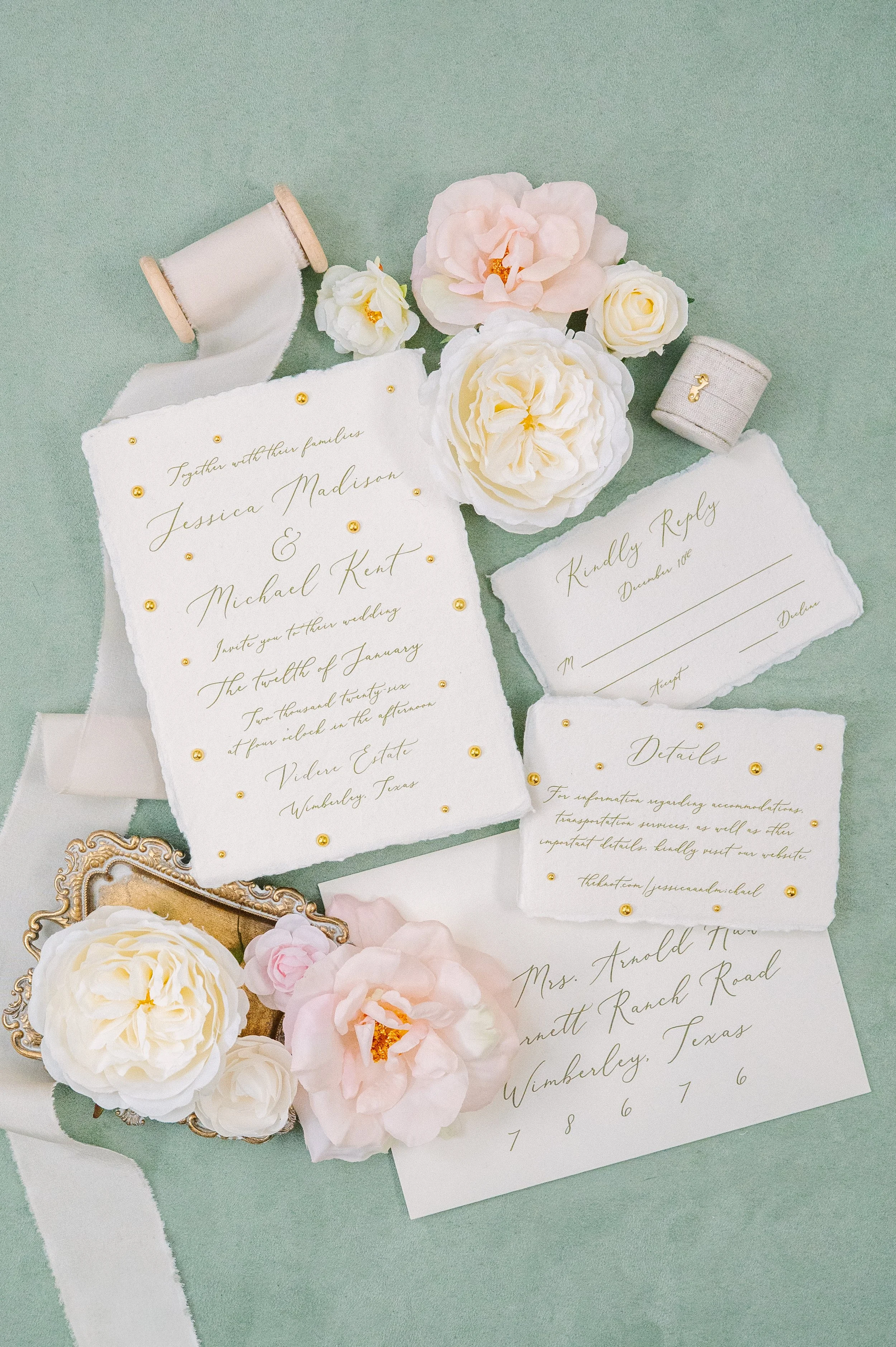 Invitation cards and reply card for a wedding, decorated with white and pink flowers, gold beads, and ribbons, laid on a green surface.