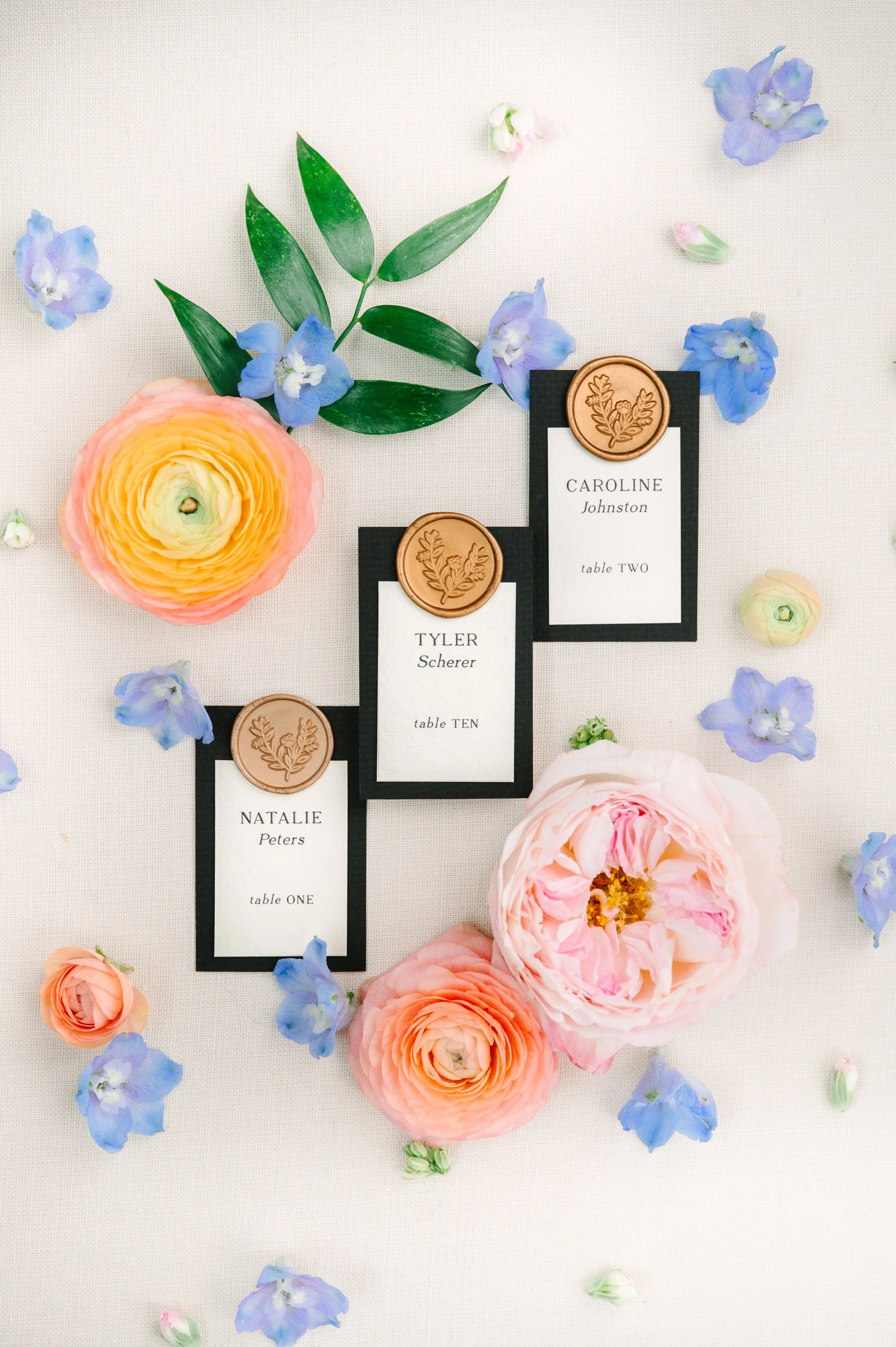 Wedding place cards with floral decorations, including pink and peach ranunculus flowers, and scattered blue flowers on a white surface.