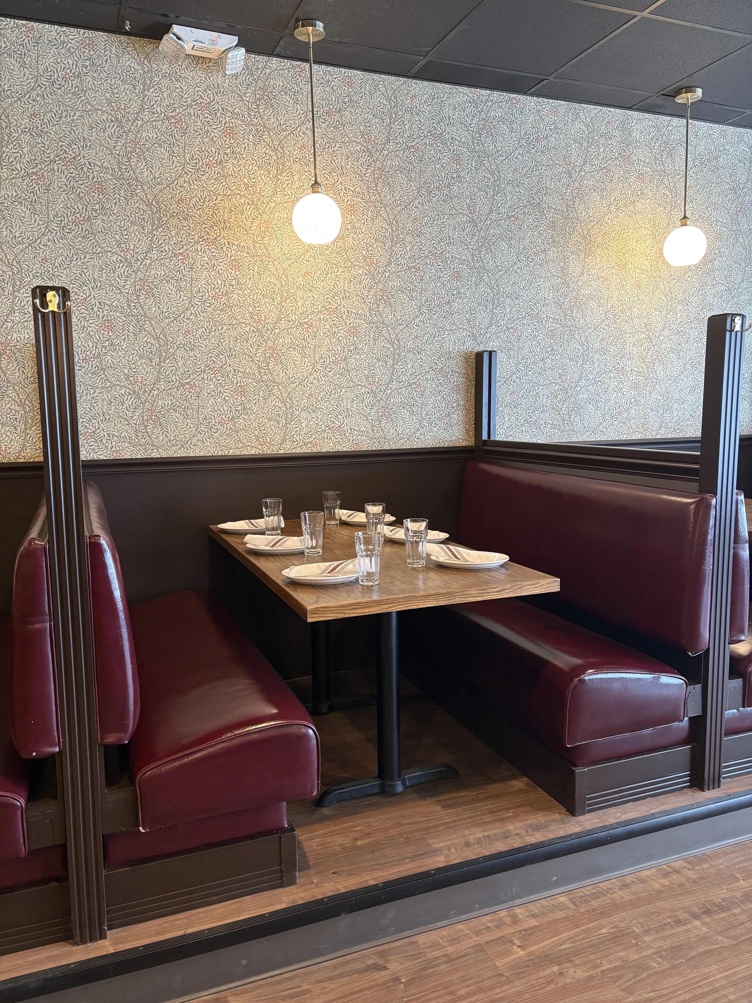 Restaurant dining booth with burgundy leather seating, set tableware, floral wallpaper, pendant lights, and mirrors