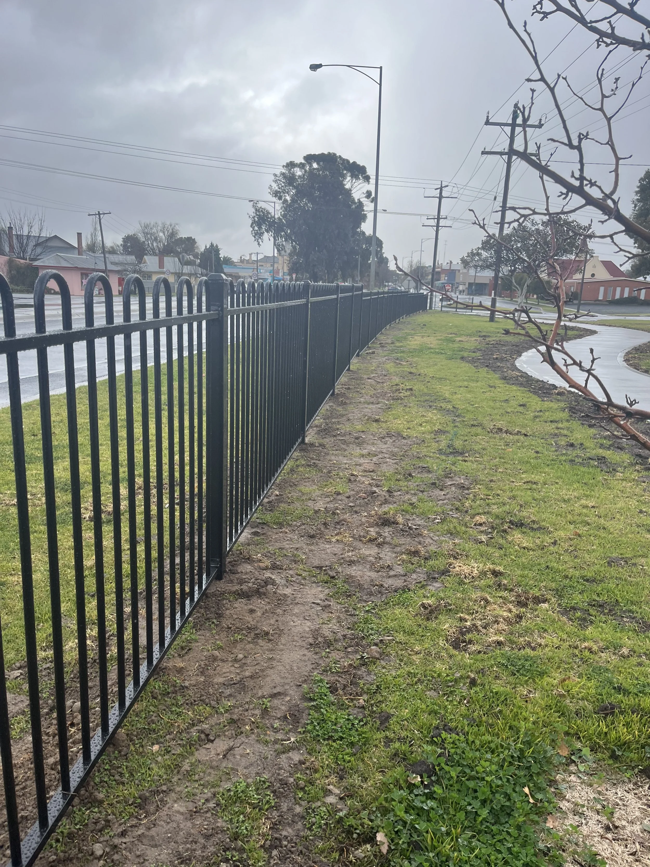 A sidewalk with a black metal fence separating it from a street, under a cloudy sky. There are patches of grass and soil along the sidewalk, with some trees and buildings in the background.