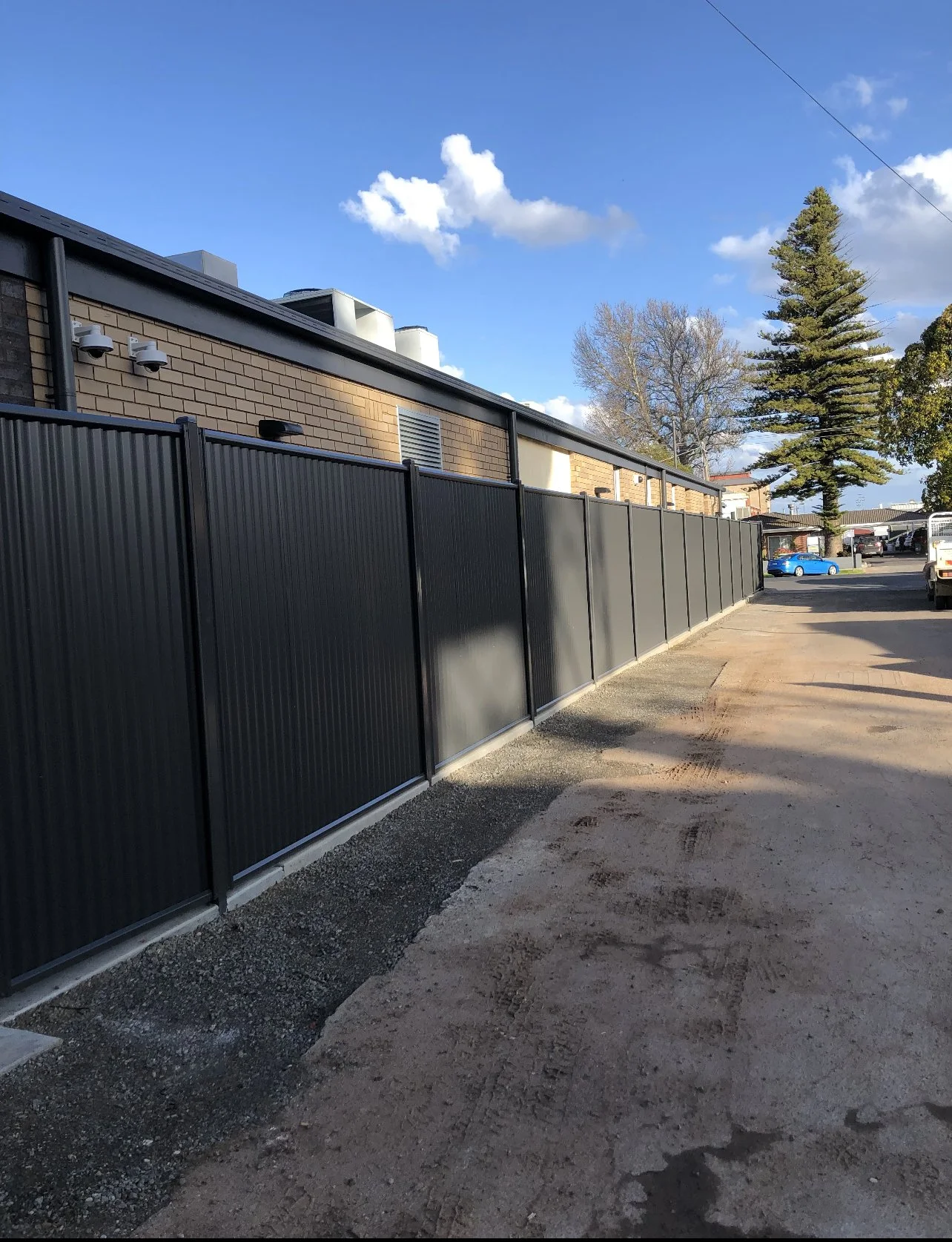 A black metal fence running along the edge of a building with a dirt and gravel pathway in front of it, under a blue sky with a few clouds and some trees in the background.