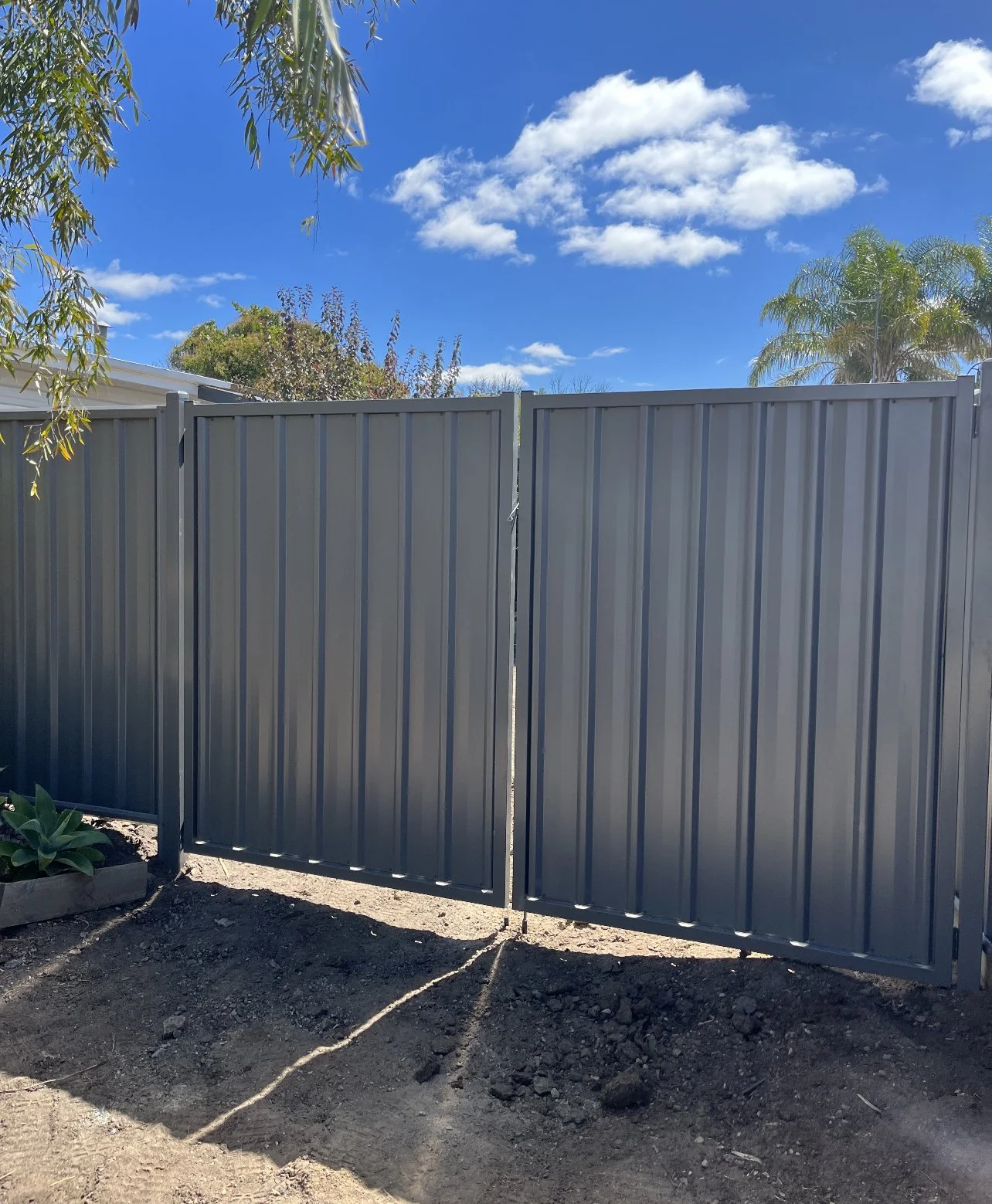 Photo of a gray metal gate under a bright blue sky with some clouds, with trees visible behind the gate.