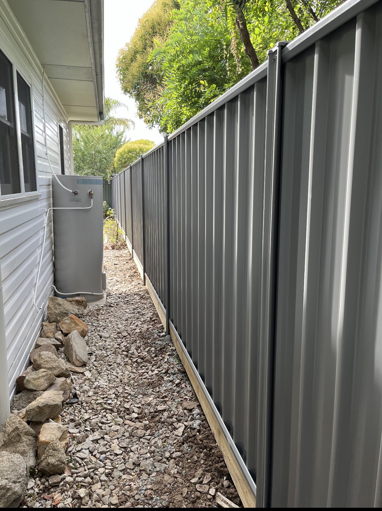 Narrow side yard next to house with gravel ground, a gray water heater on the left, and a tall gray metal fence running along the right side, with some trees and bushes visible at the end.