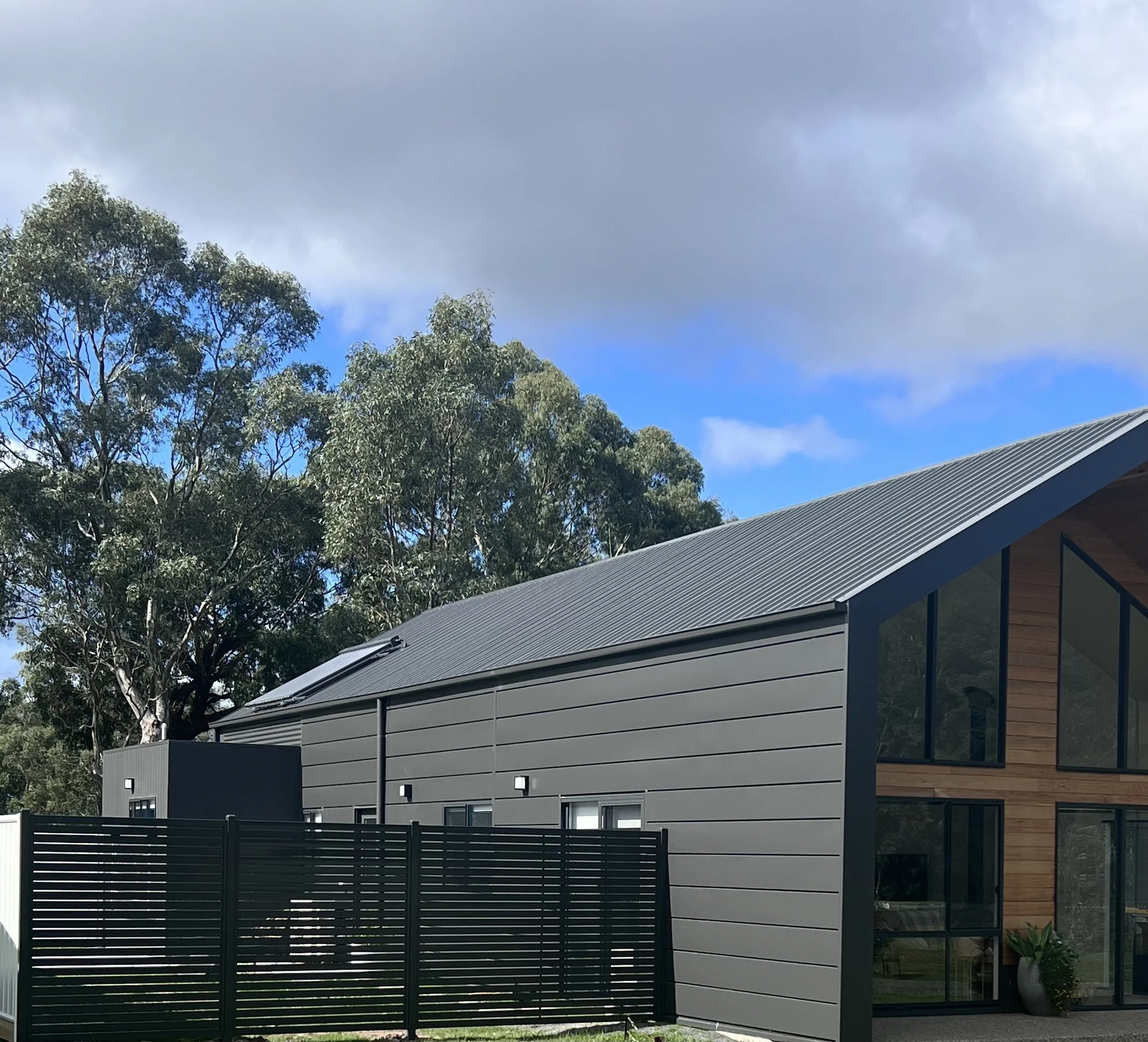 Modern house with gray siding and wooden panels, large windows, and a metal roof, surrounded by trees and a black fence.