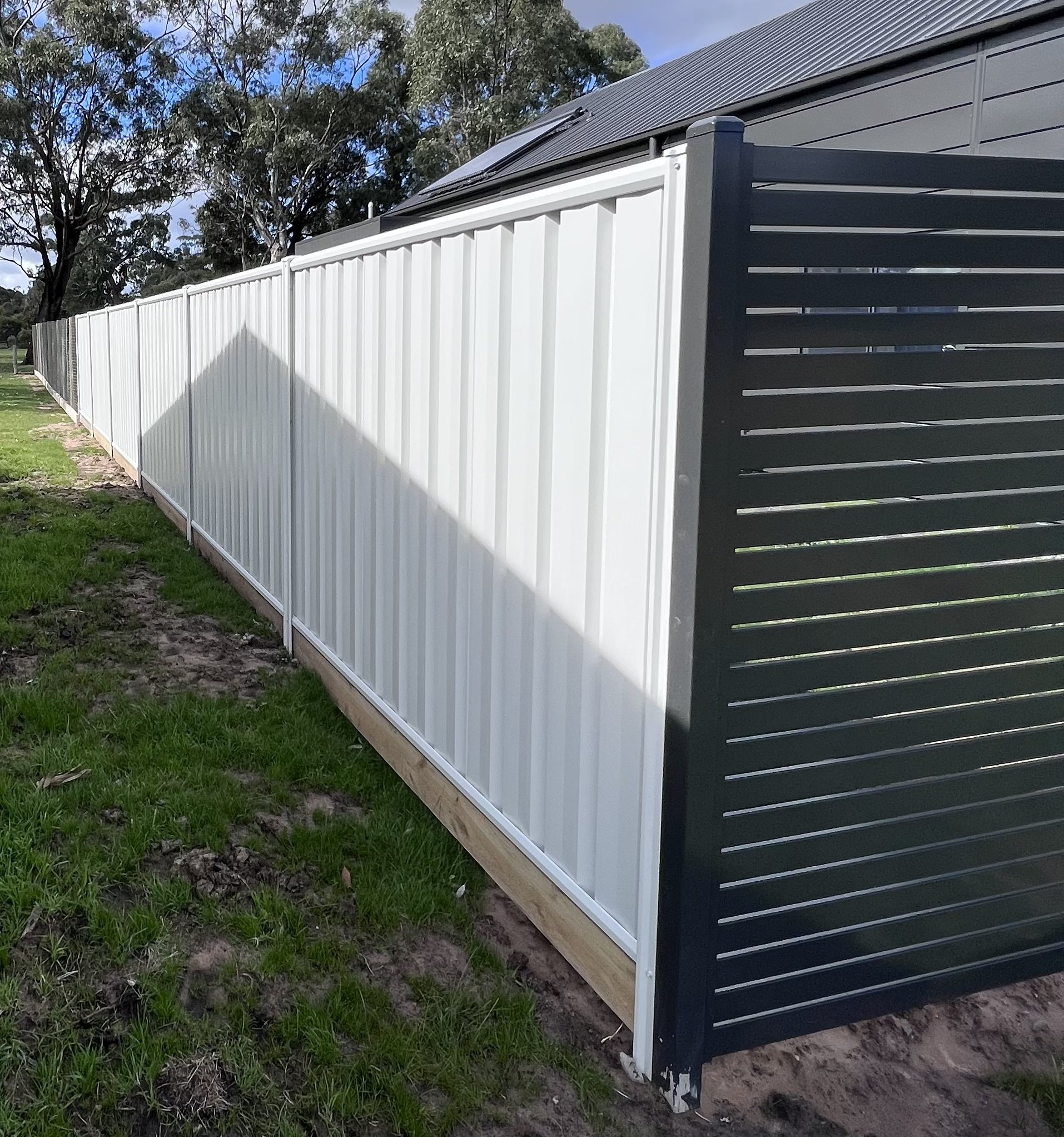 A long white fence with a black slatted door and solar panels on the roof of a nearby building, with green grass and trees in the background.