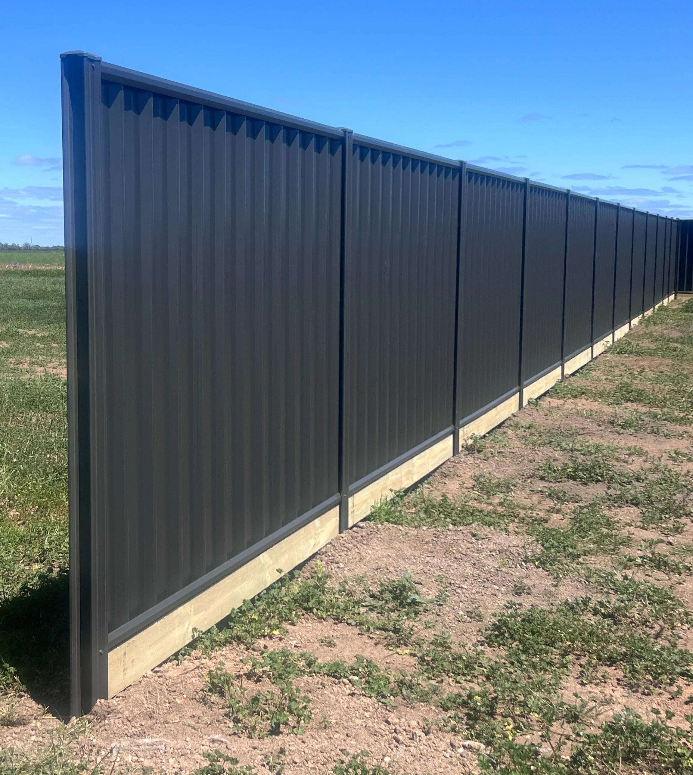 Long black metal fence with vertical slats on concrete base in an open grassy area under a blue sky with scattered clouds.