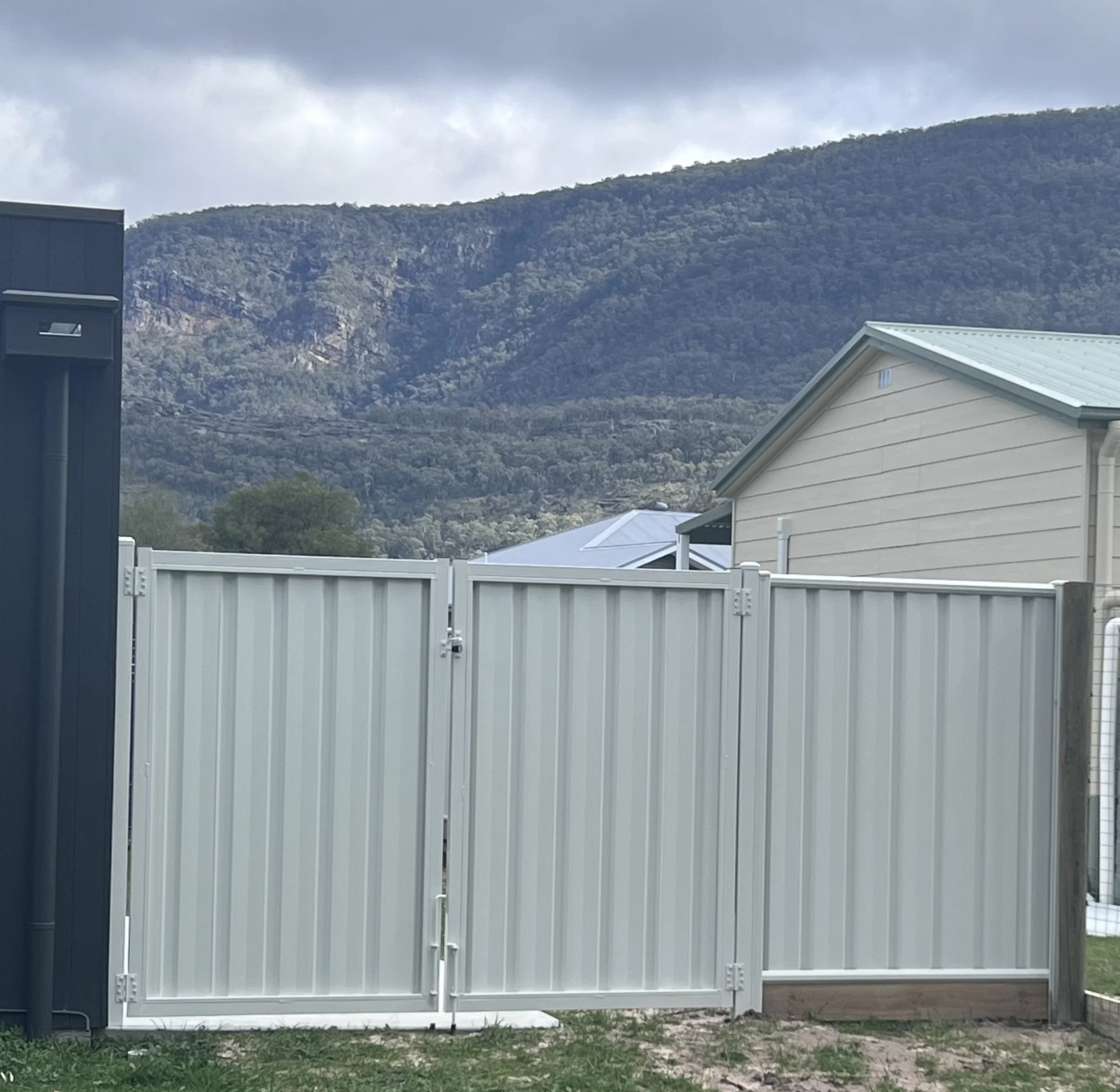 A white metal gate in front of a beige house with a green metal roof, with mountains in the background and cloudy sky above.