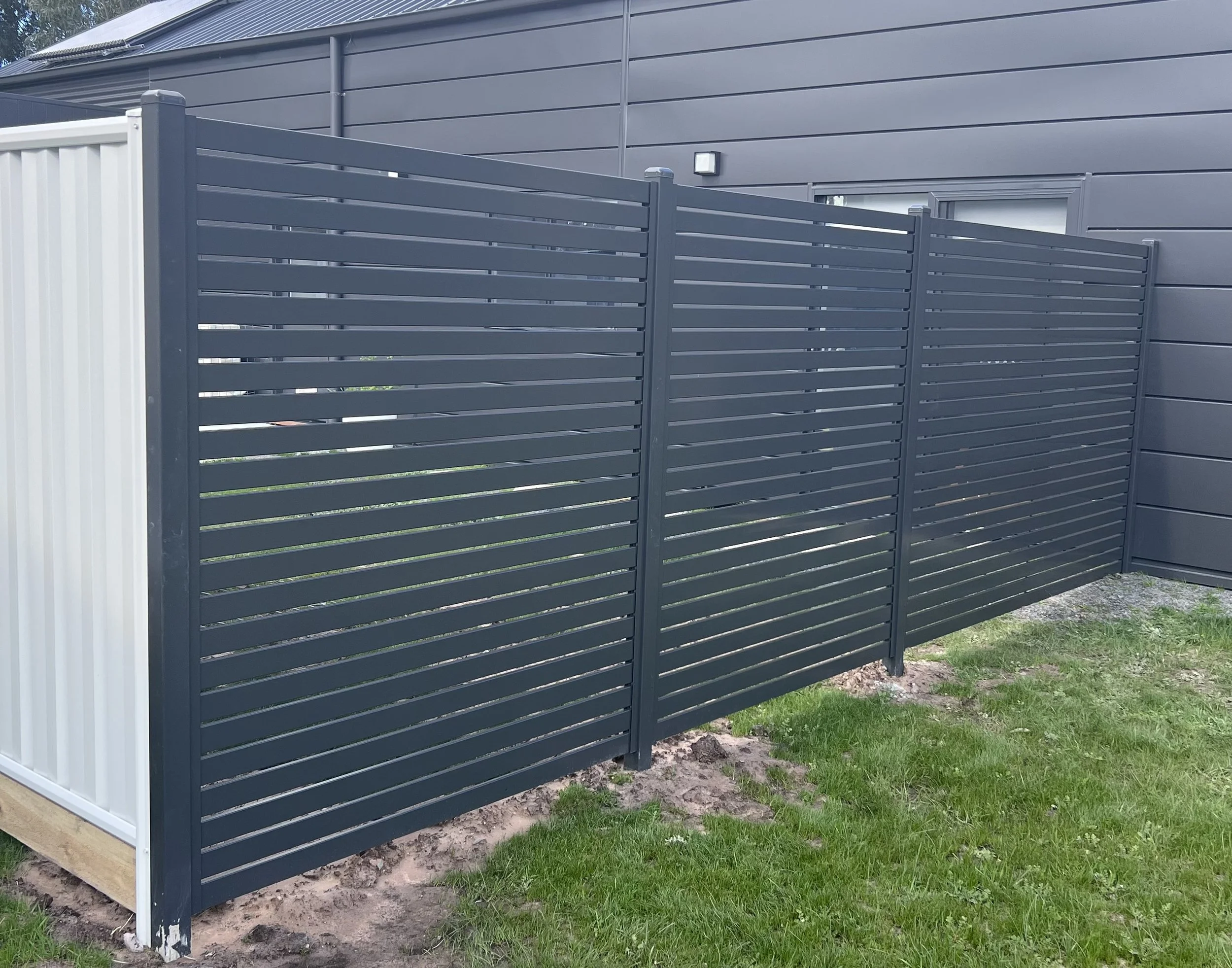 Dark gray horizontal slat privacy fence with a white section on the left, in front of a gray house exterior with a small window and a light fixture, on a grassy yard with dirt patches.