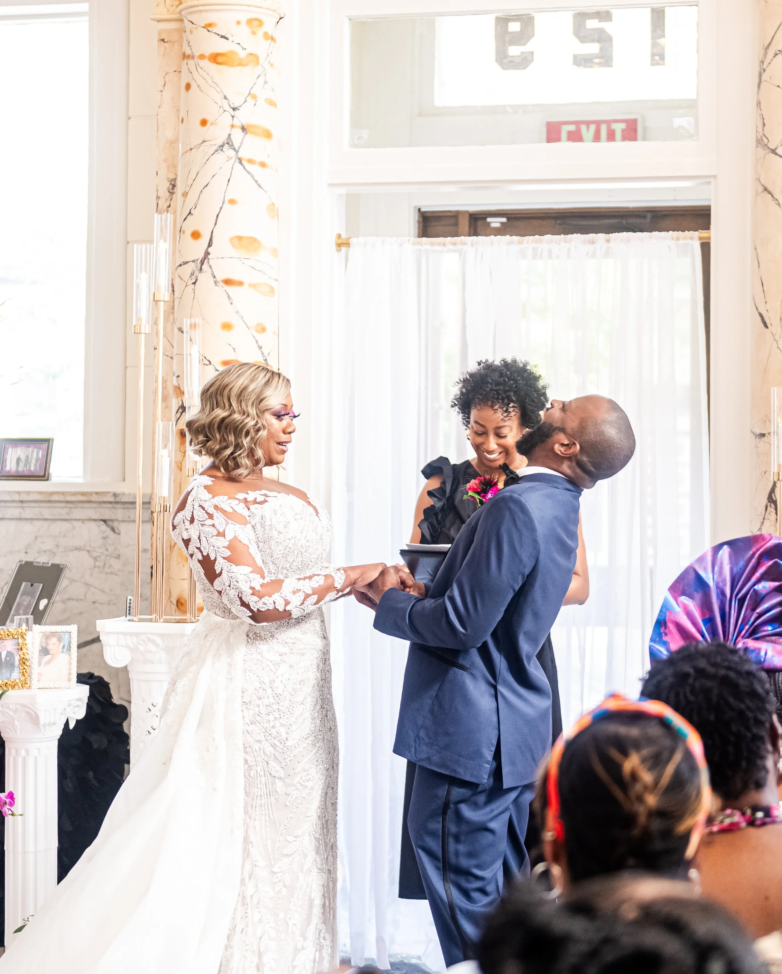 A bride and groom exchanging vows during a wedding ceremony, with a woman officiating and guests seated and watching.