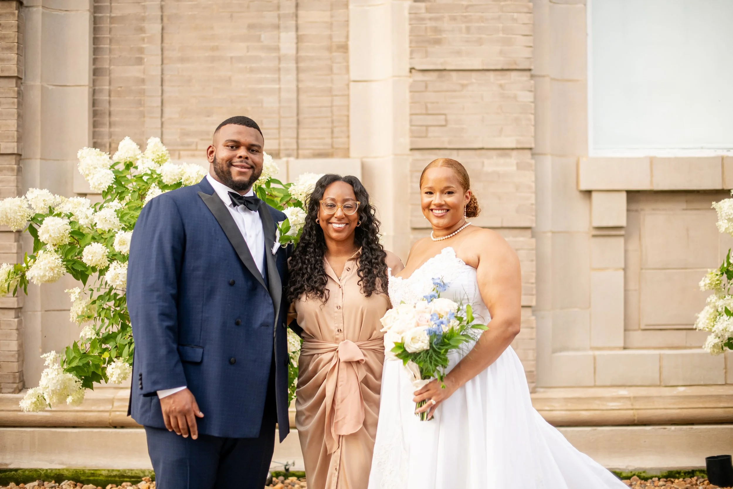 A wedding scene with a bride, groom, and a woman standing in front of a backdrop of white hydrangea flowers and brick building.