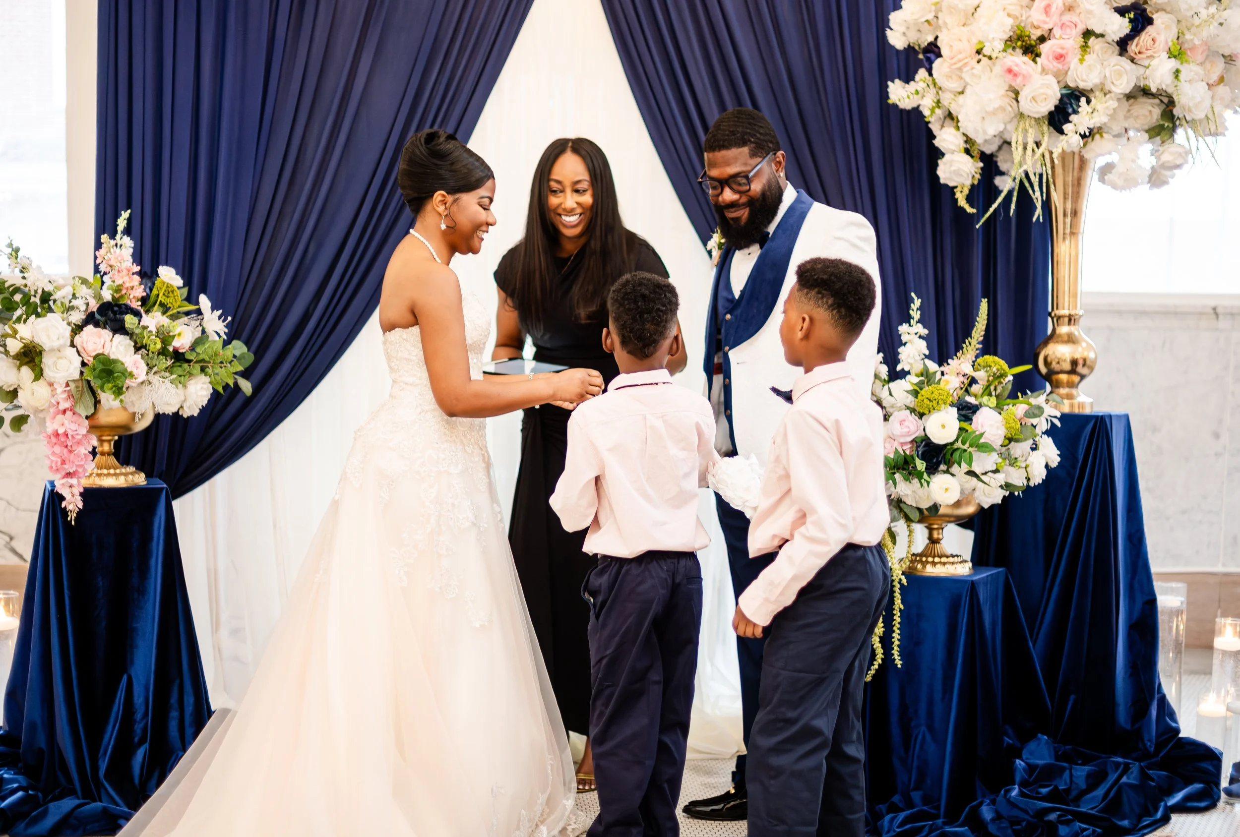 A wedding ceremony with a bride and groom exchanging rings, joined by two young boys, with a woman officiant smiling in front of a blue draped backdrop and floral arrangements.