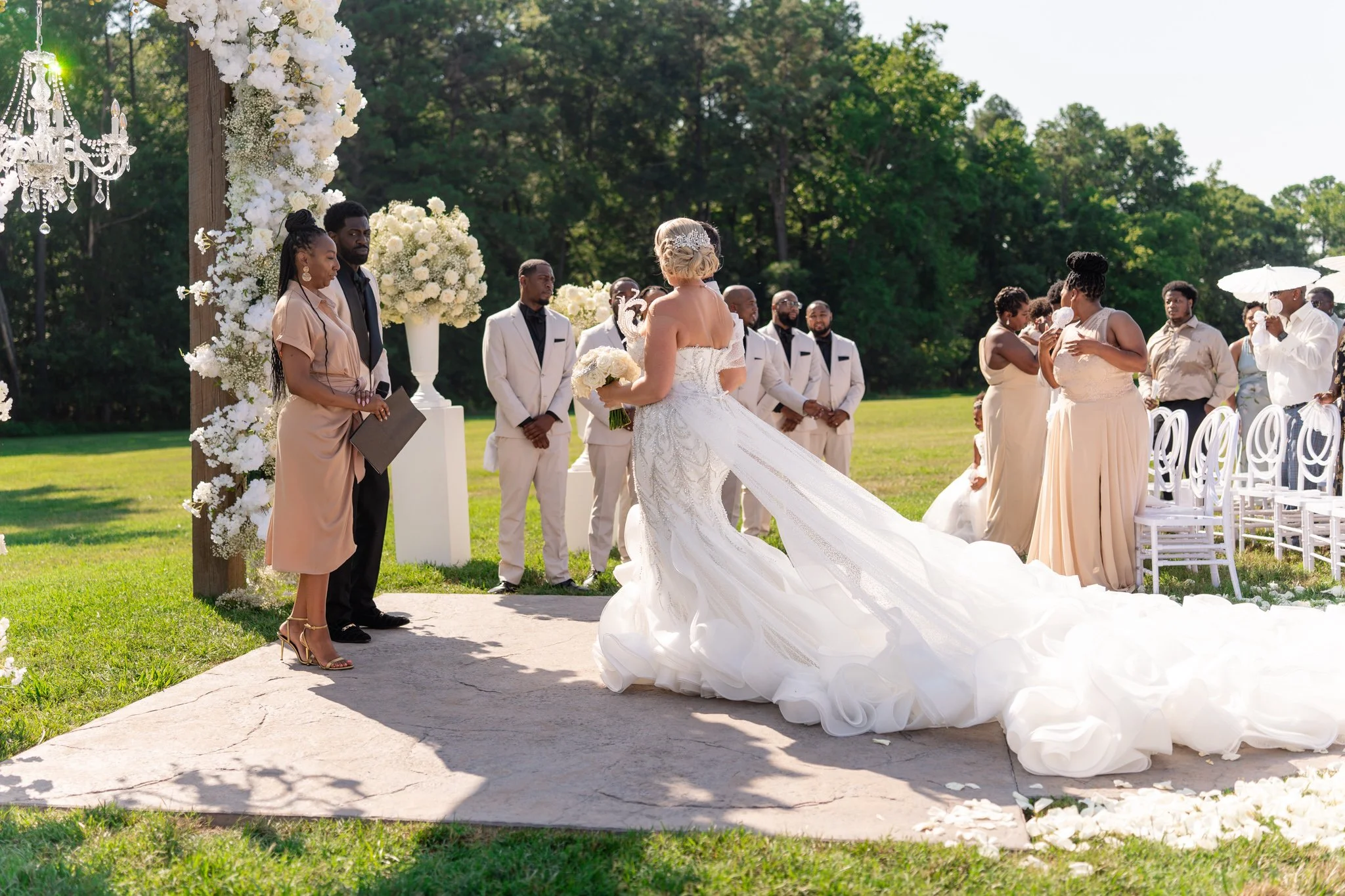 A wedding ceremony taking place outdoors on a sunny day, with a bride in a white gown and groom in white suit standing at the altar, surrounded by bridesmaids and groomsmen, with lush green trees in the background.