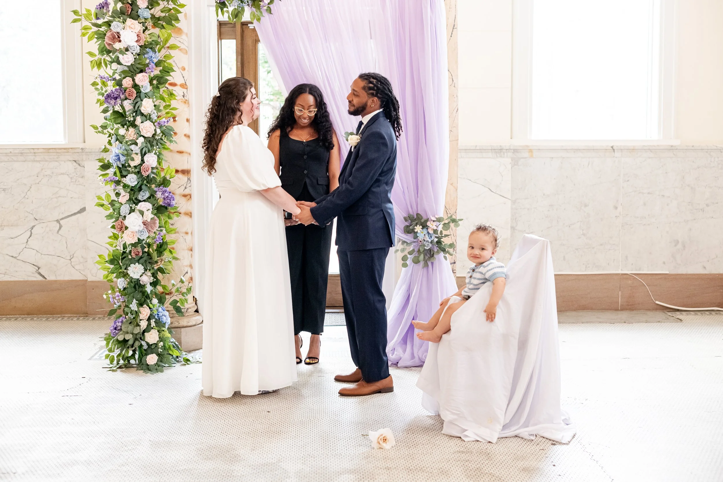 A couple is getting married in a wedding ceremony, holding hands and exchanging vows. They are standing in front of a floral arch with purple drapery. An officiant is standing behind them. A young child is seated nearby, looking towards the camera.