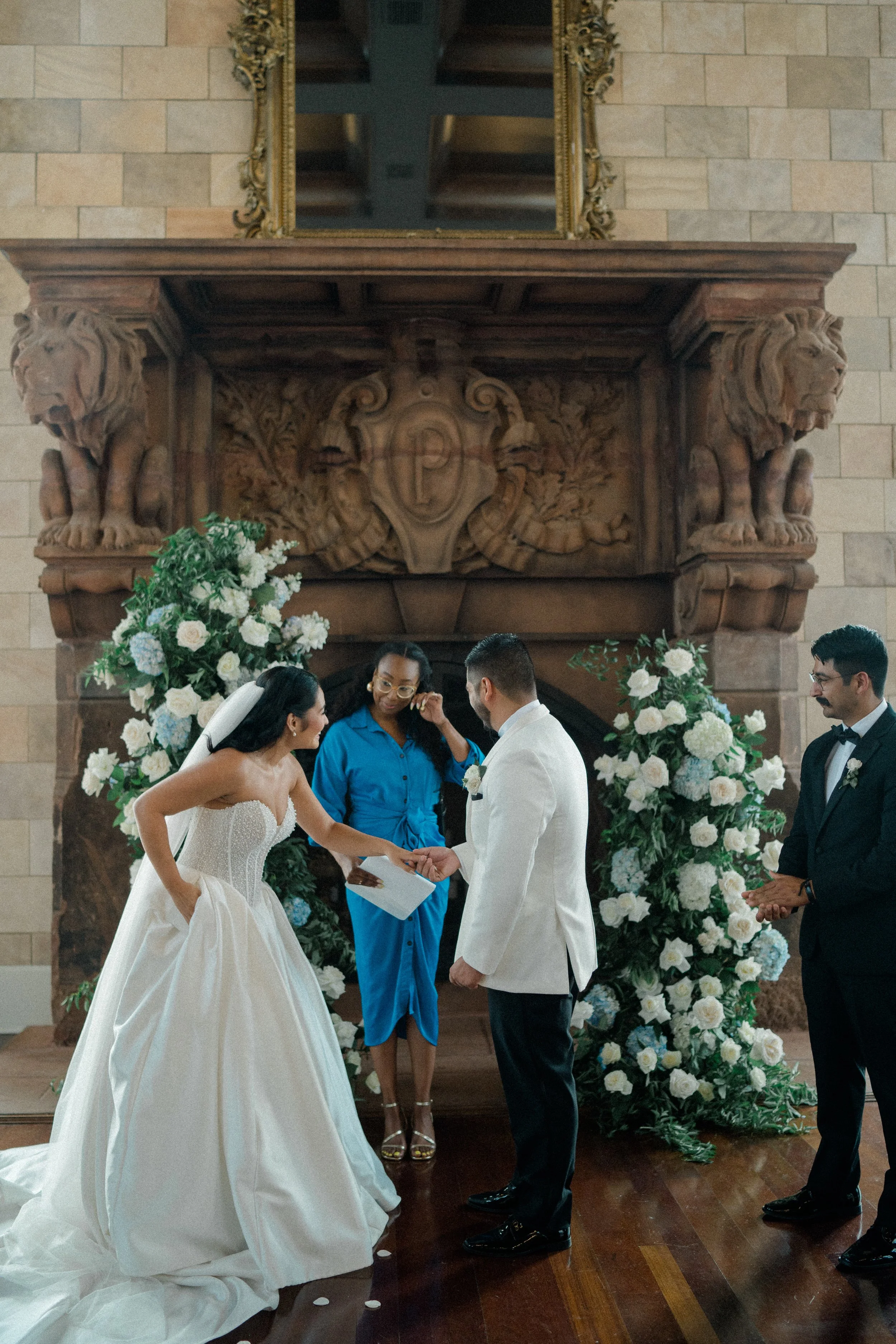 A wedding ceremony with a bride and groom exchanging vows in front of a minister, with an ornate fireplace and floral arrangements in the background.