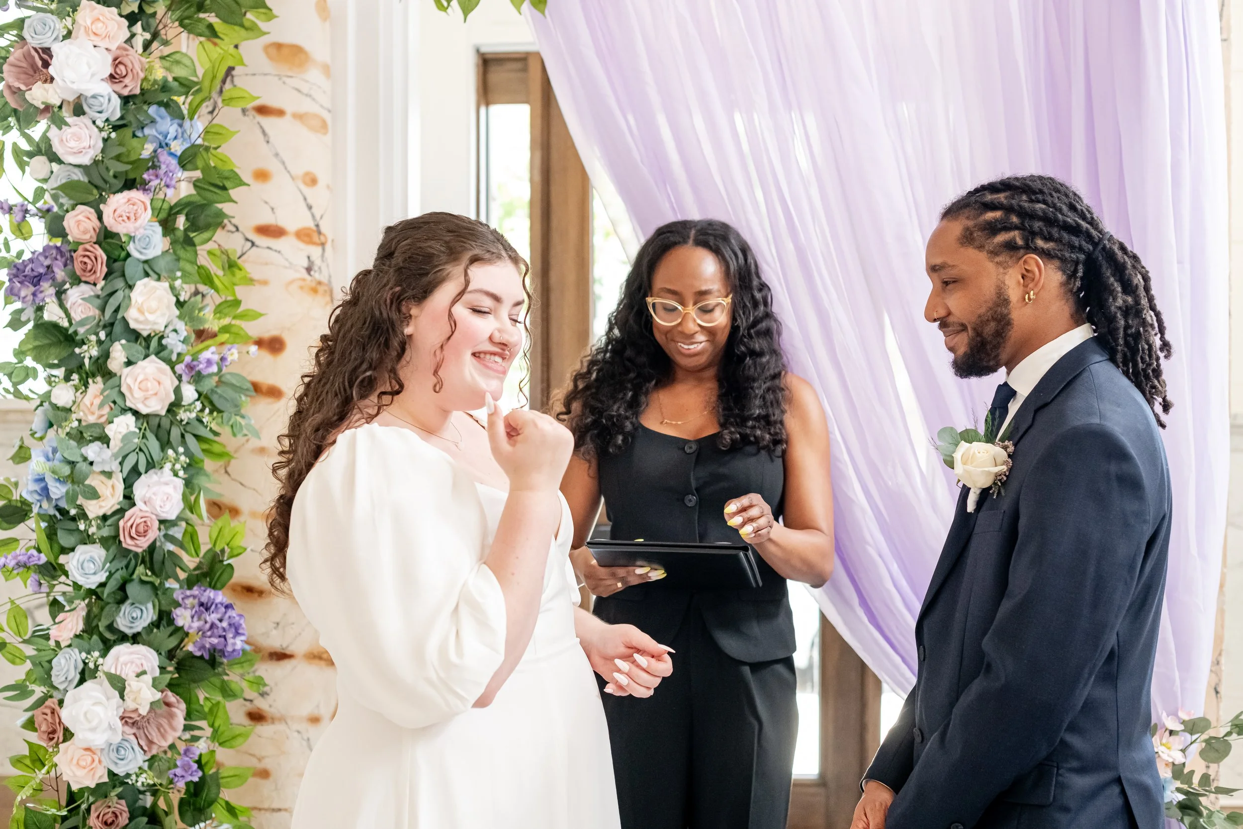 A couple getting married during the ceremony with a woman officiant standing between them, exchanging vows, with floral decorations and purple drapes in the background.
