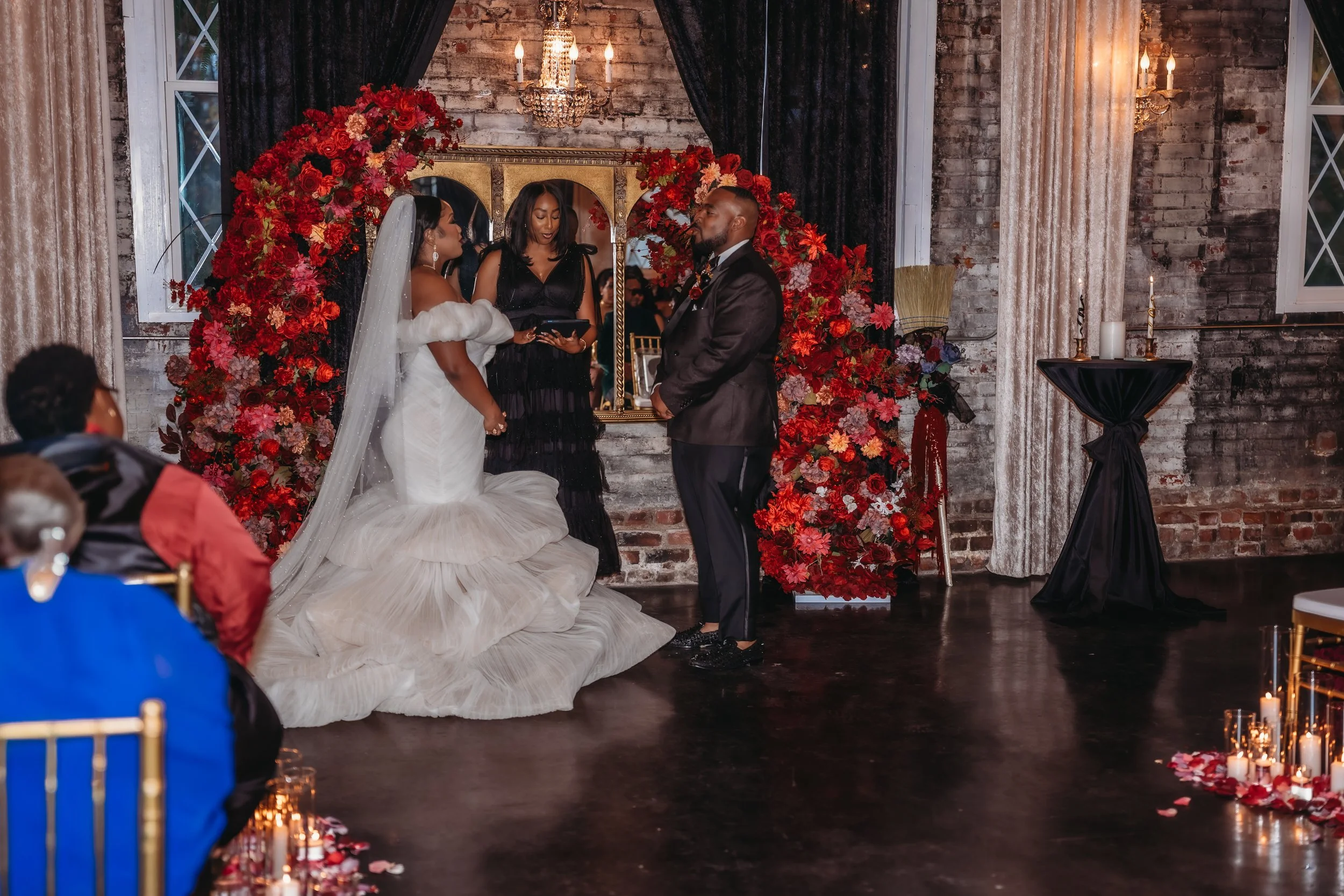 A wedding ceremony with a bride and groom standing before officiant in front of a floral arch of red and pink flowers. The bride is wearing a white wedding gown and veil, and the groom is in a black suit. The backdrop includes brick walls, black curt