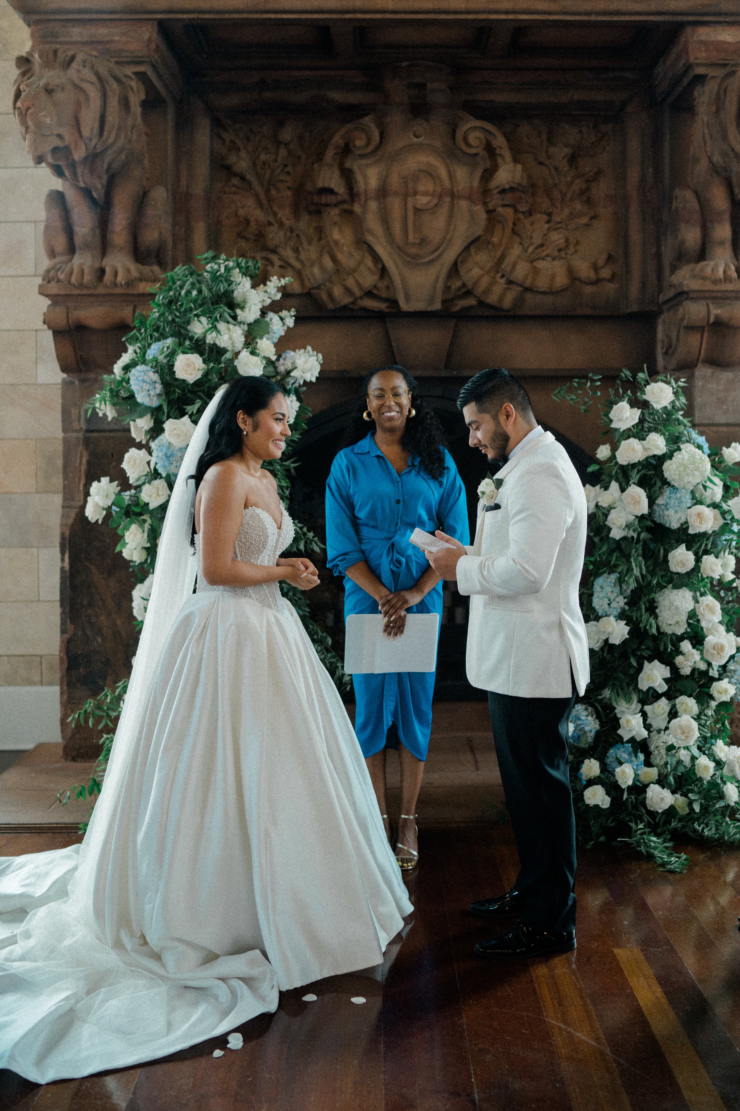 A wedding ceremony with a bride and groom standing before an officiant, surrounded by floral arrangements, in front of an ornate fireplace with two lion sculptures.
