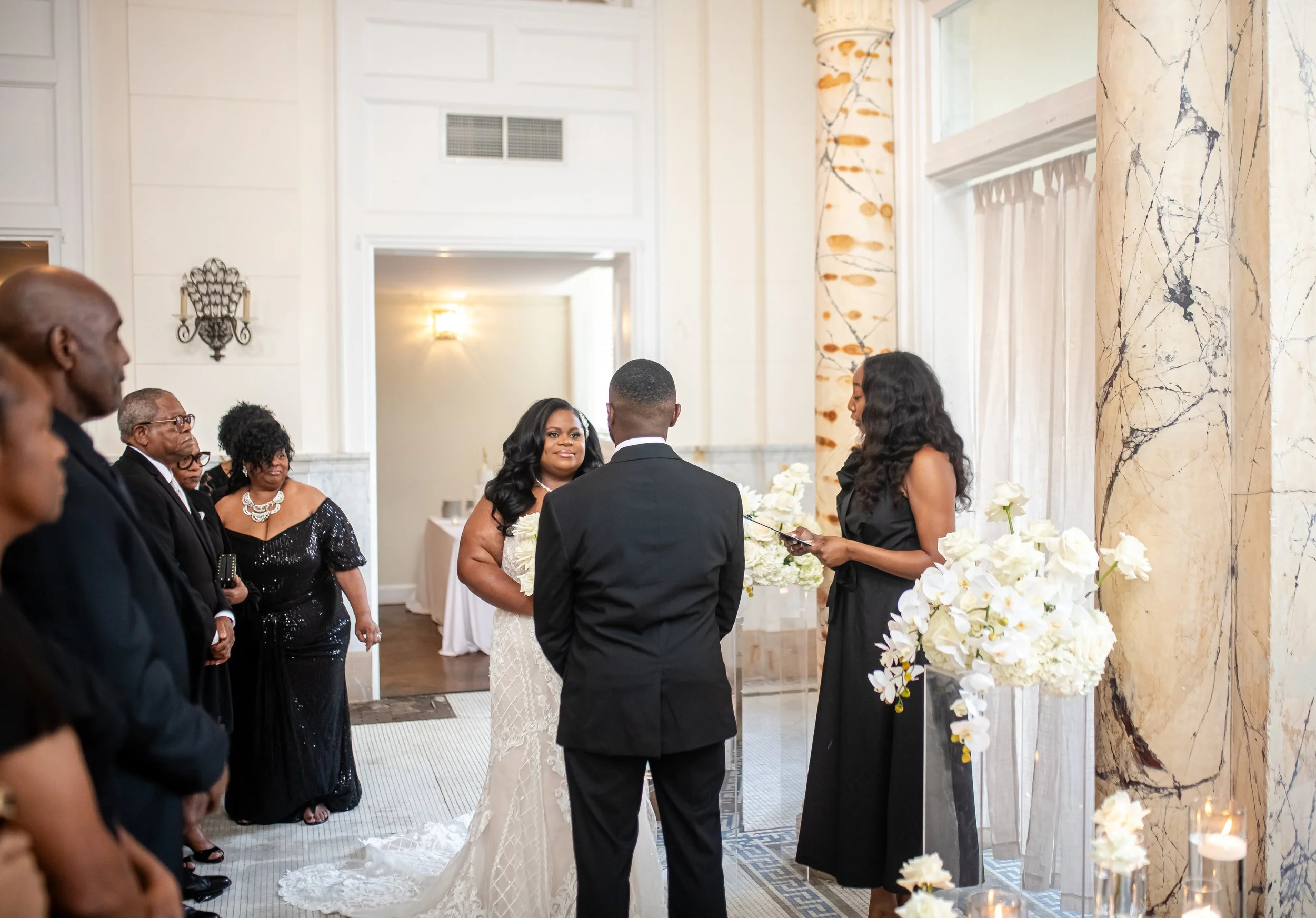 A wedding ceremony with the bride and groom standing facing each other, surrounded by family and friends in a decorated indoor space with white flowers and elegant decor.