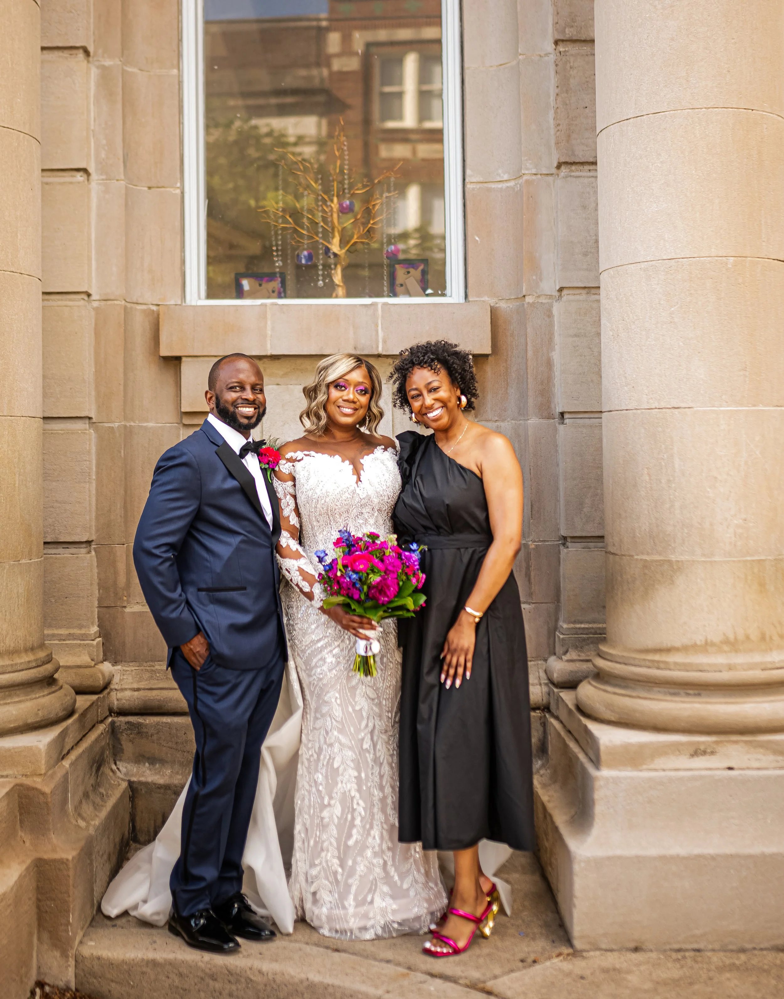 Three people standing outside a building with stone columns, dressed in formal attire for a wedding. The woman in the middle is in a white bridal gown, holding a bouquet of pink, purple, and green flowers. The man on the left is in a navy tuxedo, and
