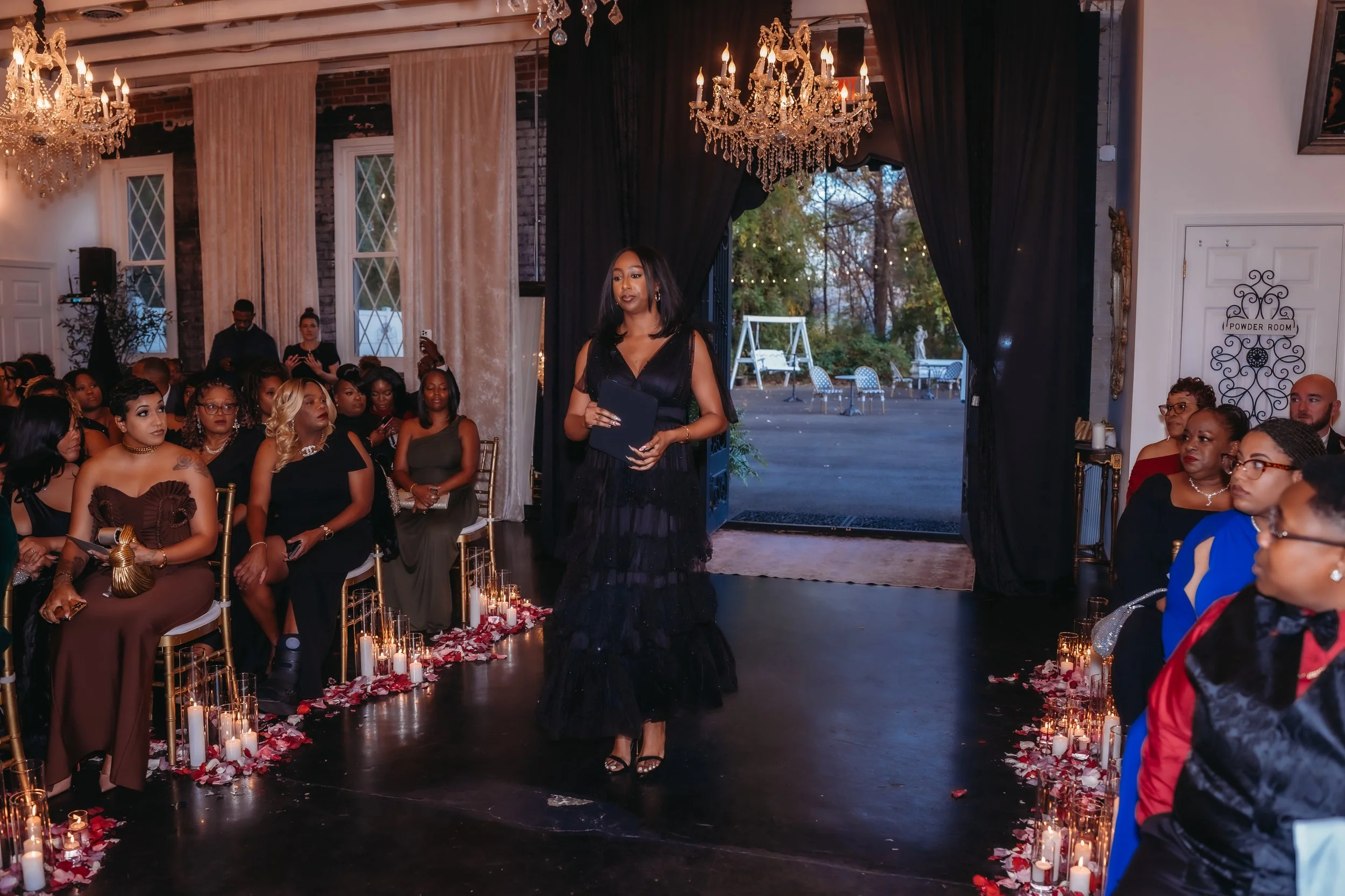 A woman in a black dress walking down an aisle decorated with candles and rose petals, during an indoor event with seated audience members.