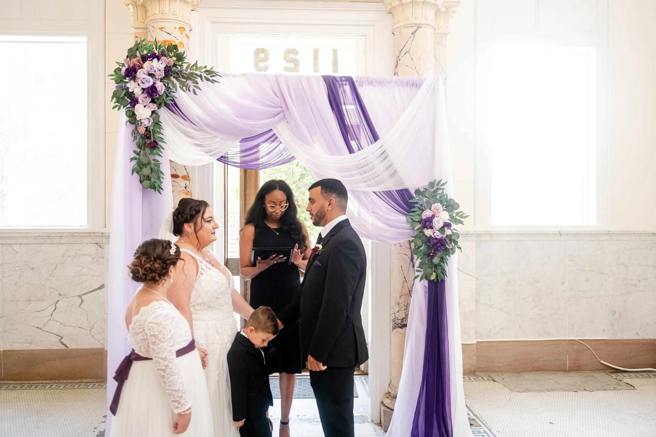 A wedding ceremony with a bride, groom, two women, and a young boy standing under a purple and white floral wedding arch inside a marble-walled building.