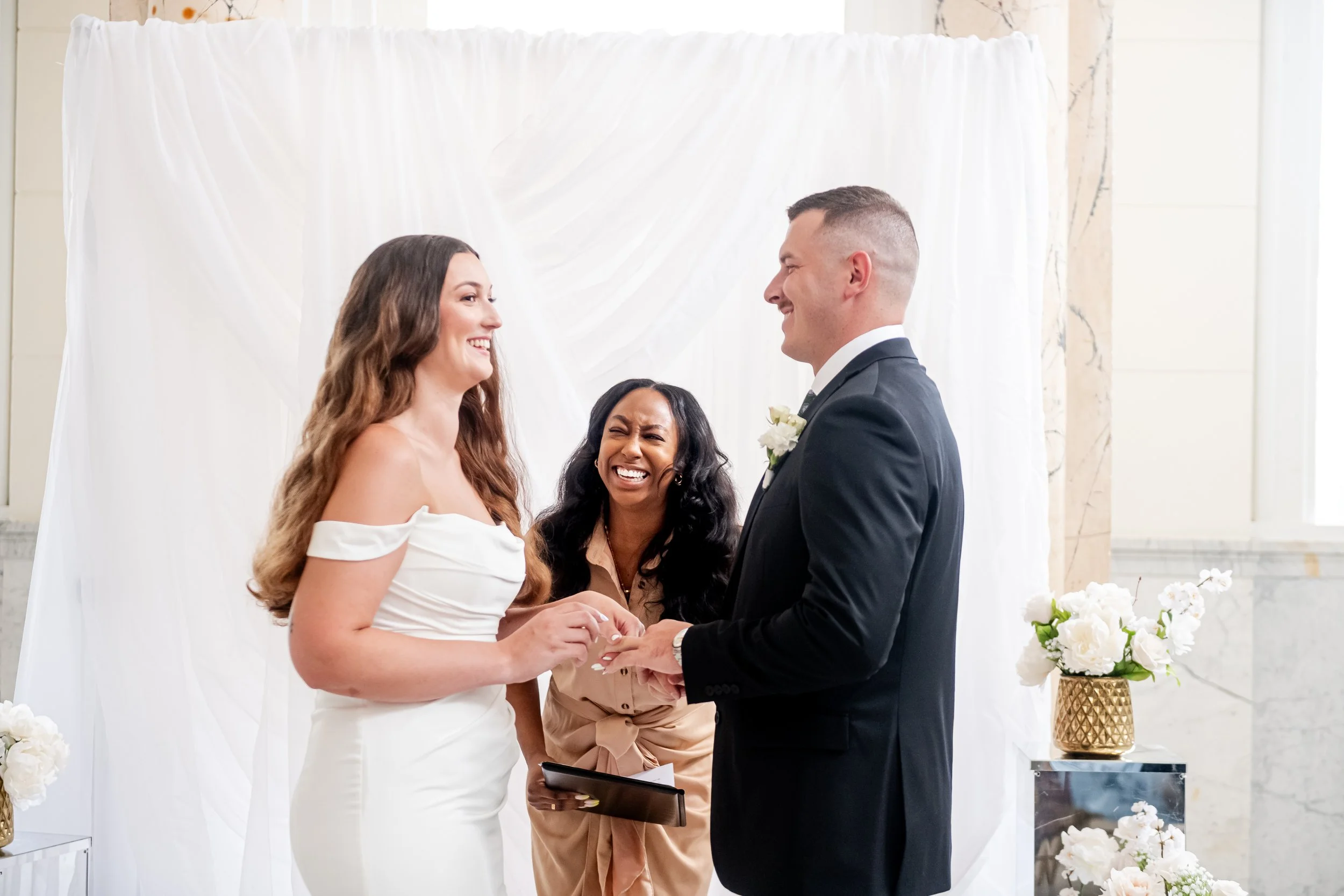 A bride and groom exchanging wedding rings during a wedding ceremony, with a officiant standing behind them, all smiling and happy.