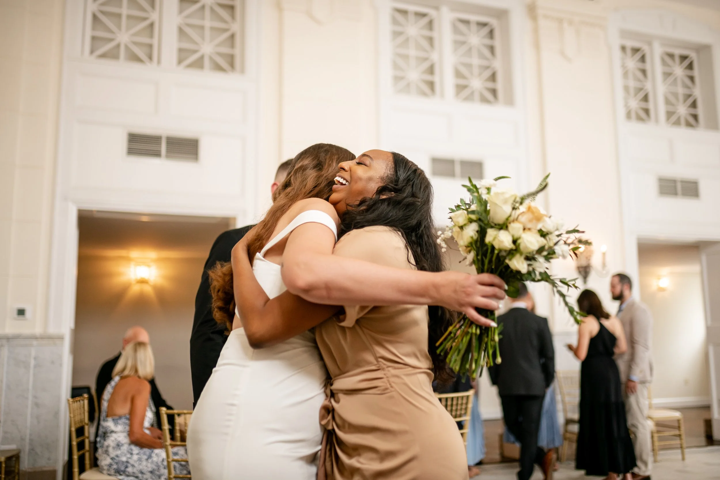 Two women hug at a wedding reception, one holding a bouquet of white flowers, in an elegant room with high ceilings and ornate decor.